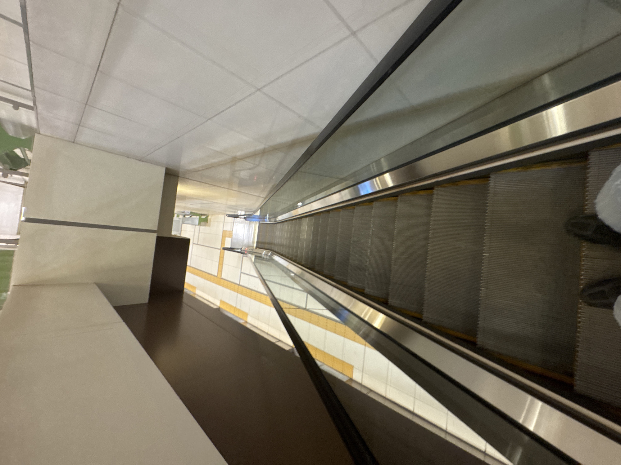 The image shows a downward-facing view of an escalator in what appears to be a modern public space, possibly a shopping mall or transportation hub, in Sofia, Bulgaria. The escalator's metallic steps are clearly visible, descending into a dimly lit area. The sides of the escalator feature glass panels with metallic handrails, reflecting the surrounding environment. To the left, a dark brown wall contrasts with a lighter, cream-colored wall panel above. Yellow tactile paving is visible on the floor to the left of the escalator, indicating a pathway. The overall environment is clean and functional, with the lighting casting shadows and highlighting the textures of the materials. Two pairs of shoes are visible at the bottom of the frame, suggesting the presence of at least two individuals on the escalator.