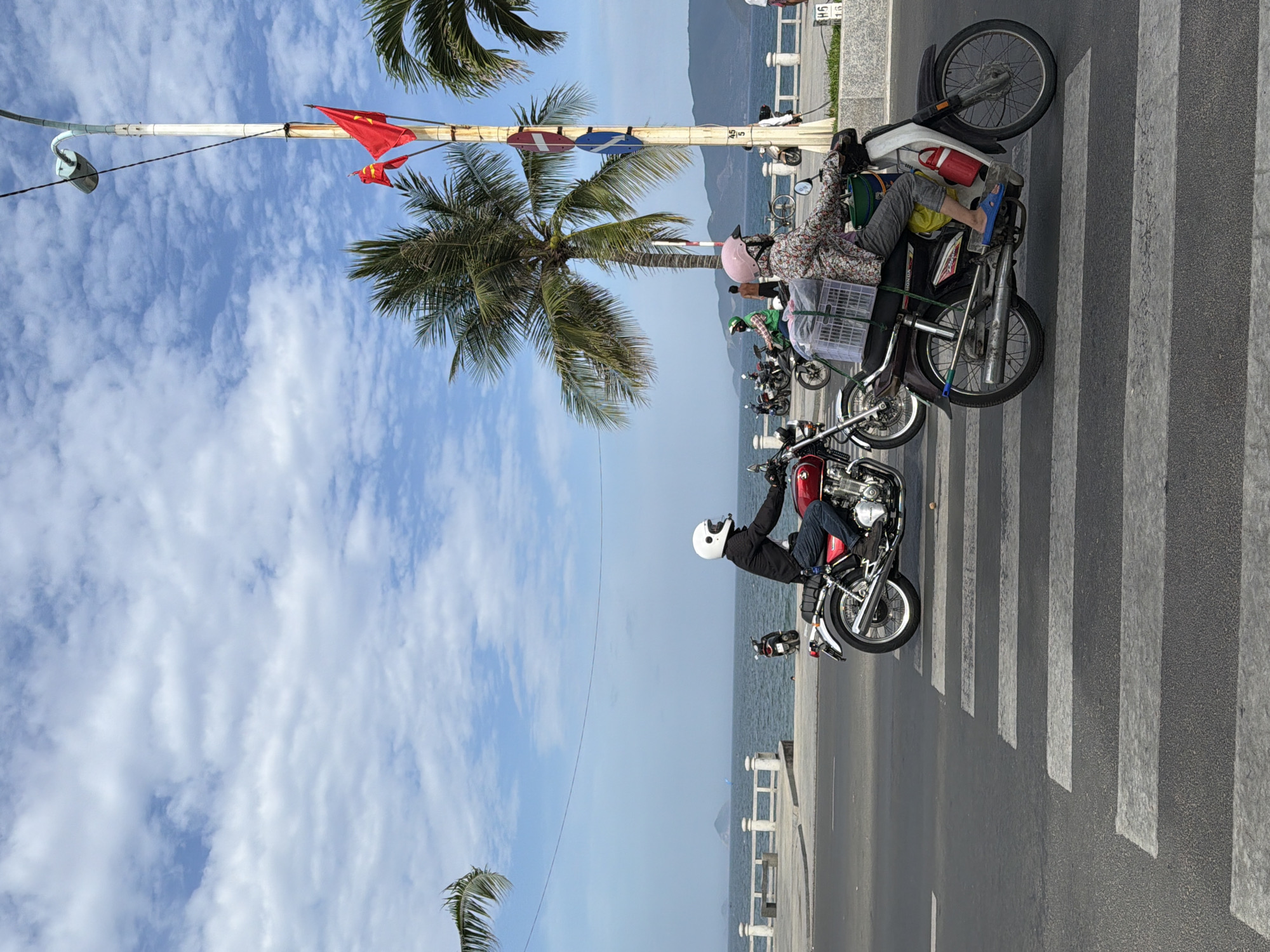 The image depicts a sunny day in Nha Trang, Vietnam, likely along the coastline, given the presence of the sea and palm trees. Two individuals are riding motorbikes on a road that has a pedestrian crossing in the foreground. The motorbike on the right is ridden by a person wearing a floral shirt, grey pants, a pink helmet, and blue sandals, and the motorbike is carrying a large white plastic crate secured to the back. The motorbike on the left is ridden by a person wearing a black jacket, blue jeans, and a white helmet. In the background, several other motorbikes are visible, as well as a white railing along the promenade, palm trees, the sea, and distant mountains. A tall lamppost with two red flags is also present. The sky is blue with scattered white clouds. The road appears to be a main thoroughfare in the city.