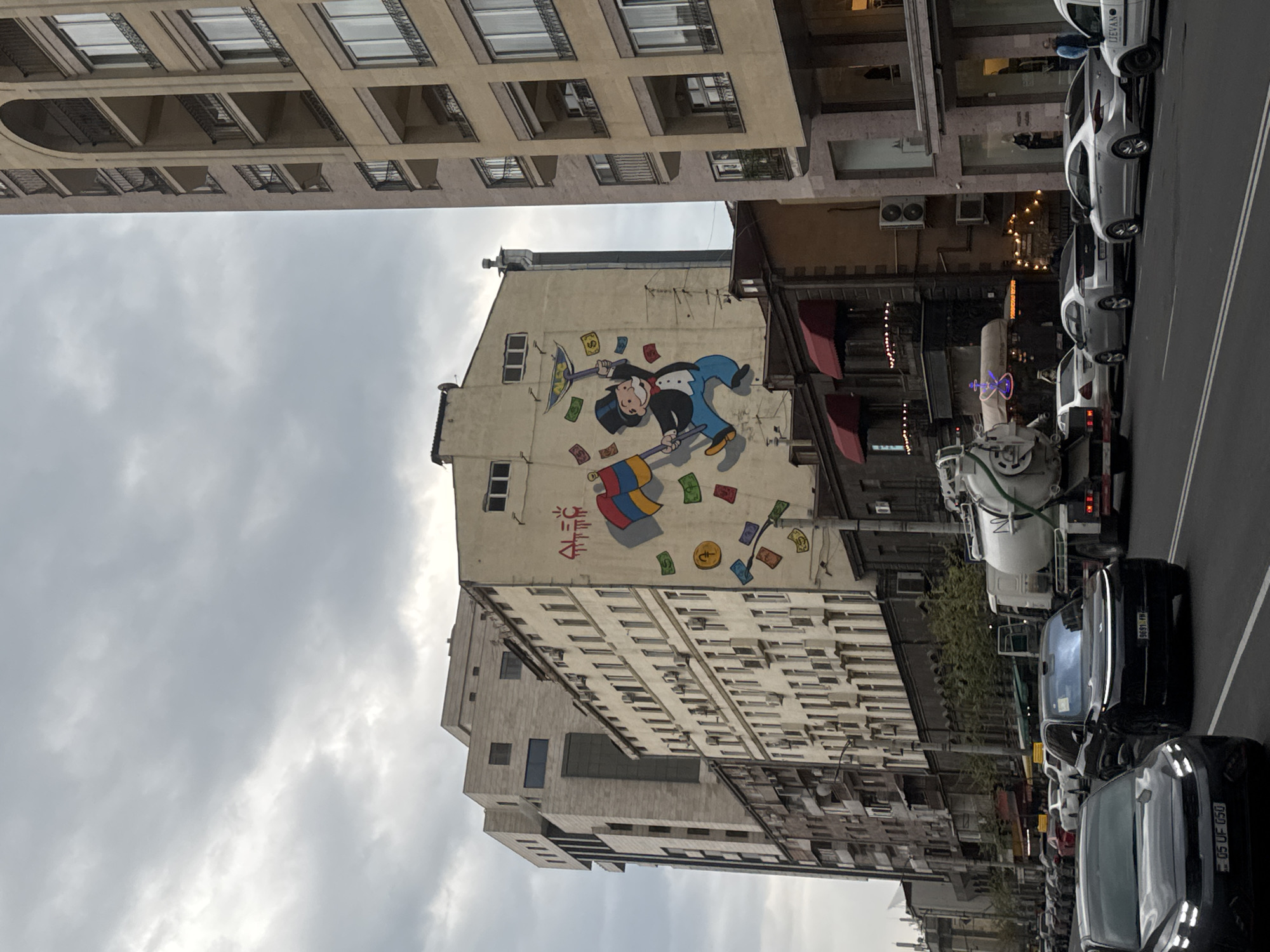 This rotated image captures a dynamic street scene in Yerevan, Armenia, under a cloudy sky. The main focal point is a large mural on the side of a light-colored building, which is positioned prominently in the center of the frame.
The mural depicts a cartoon character resembling Mr. Monopoly (Rich Uncle Pennybags), dressed in his characteristic top hat, suit, and monocle. He is shown actively gathering currency: using an open umbrella to catch falling dollar bills and a stick to sweep more bills and gold coins. The money symbols include prominent dollar signs. Next to the character, red text in Armenian script is visible, which appears to be "ՀԱՂԹԱ" (Haghta), meaning "WIN" or "VICTORY". Below the Monopoly figure, another colorful, abstract graphic resembling a segmented umbrella or fan is depicted, also surrounded by currency.
To the left of the mural building, a taller, modern building with multiple stories, numerous windows, and balconies with metal railings extends upwards, partially obscured by the cloudy sky.
The right side of the image features a street lined with parked cars, indicating an urban environment. Several modern sedans are visible, and their Armenian license plates, displaying "AM" and regional codes like "06" and "05" (Yerevan), confirm the location. A large white utility truck, possibly a cement mixer, is also parked on the street. One white car explicitly bears the text "YEREVAN" on its side. The time of day is daytime, but the overcast sky suggests muted light. Air conditioning units are visible on some of the buildings.