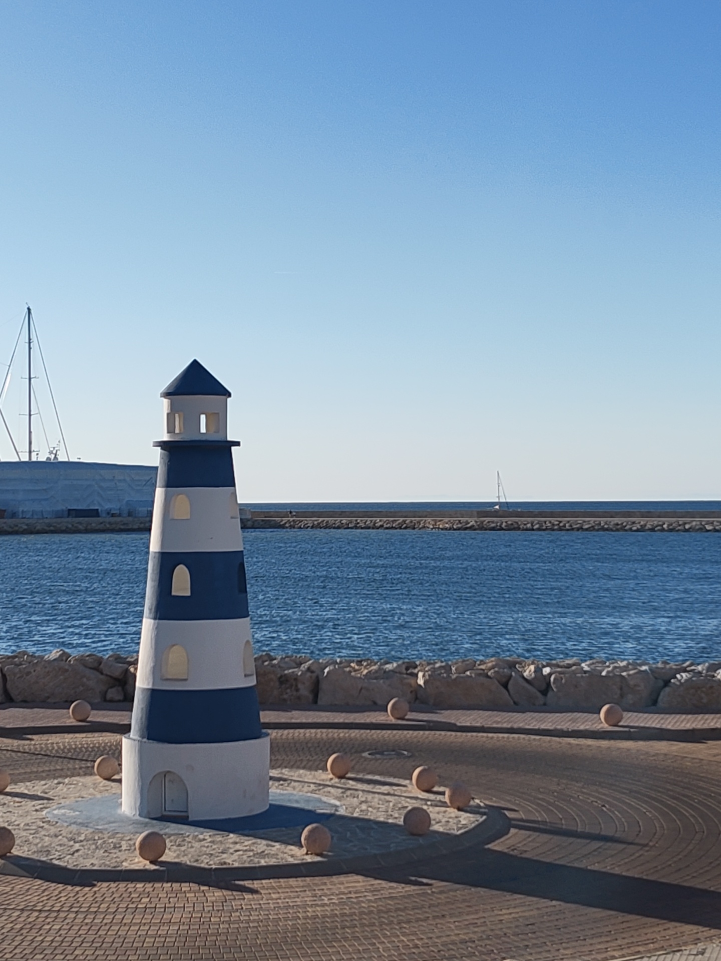 The image depicts a small, striped lighthouse replica in Dénia, Spain. The lighthouse, painted in blue and white bands, stands on a circular platform decorated with light brown, spherical bollards and a concentric brick pattern. Behind the lighthouse is a stone breakwater protecting the harbor. The water is a deep blue, with a distant sailboat visible. In the background, to the left, is a large, covered structure possibly related to the marina. The sky is a clear, bright blue. The overall scene suggests a sunny day at the harbor.