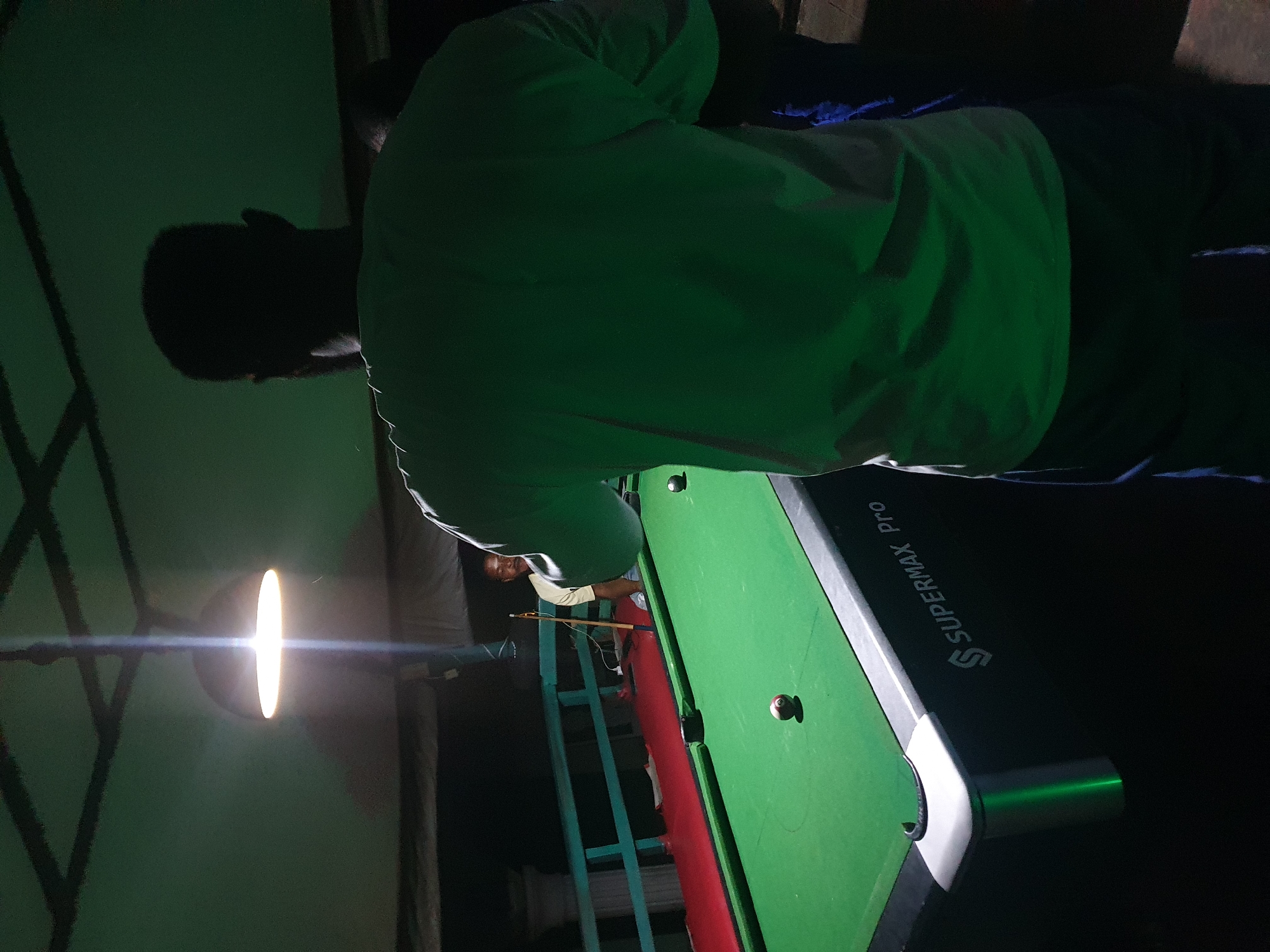 Two individuals are engaged in a game of pool. The scene appears to be an outdoor, covered area, possibly a patio or veranda, in Lokoja, Nigeria. The lighting suggests it is evening or nighttime, with a single overhead lamp illuminating the pool table. The table itself is green felt, with several billiard balls visible, including a cue ball and a striped ball. One person, seen from behind, is wearing a light-colored t-shirt and dark trousers, standing over the pool table. The other person is visible in the background, standing behind the table, wearing a light-colored t-shirt and blue trousers. A pool cue rests against the table. The "SUPERMAX Pro" logo is visible on the side of the pool table. The surrounding environment is dark, with hints of railings and possibly structures in the background.