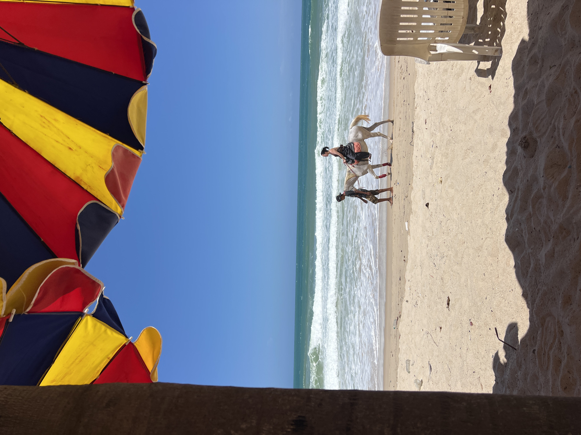 In the city of Choeng Thale, Thailand, a woman rides a white horse along a sandy beach, accompanied by a man walking beside her. The ocean waves are rolling in, creating a foamy surf. In the foreground, a portion of a large, colorful umbrella with red, yellow, and blue panels is visible, along with a palm tree trunk and a plastic chair. The sky is clear and blue.