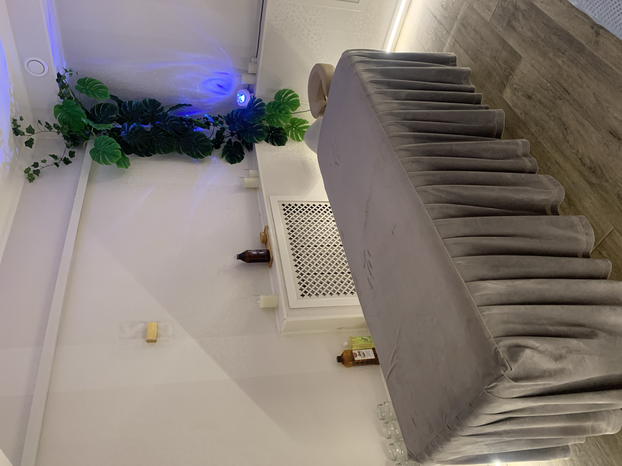 The image shows an indoor scene, likely a hallway or staircase, in Almaty, Kazakhstan. A prominent feature is a gray, fabric-covered staircase that occupies a significant portion of the frame. The staircase appears to be unusually wide and has a soft, plush texture with a decorative, ruffled edge along each step. To the left of the staircase, a white wall is adorned with artificial green foliage, including large monstera leaves and smaller ivy strands. A blue light source casts an ethereal glow on the ceiling and the plants.
On the wall near the plants, several small white candles are visible, along with a decorative wooden shelf holding a dark glass bottle. Below this, a white ventilation grate is mounted on the wall. Further down, on another shelf, are more bottles, including a clear bottle with green liquid and a brown bottle with dark contents. A few small glass jars are arranged in a row near the bottom right of the frame. The floor is covered with wood-look laminate flooring, laid in a diagonal pattern. A strip light runs along the top of the wall on the right, providing ambient lighting. The overall impression is of a modern, stylish interior with a touch of nature and soft lighting. No people are visible in the image.
