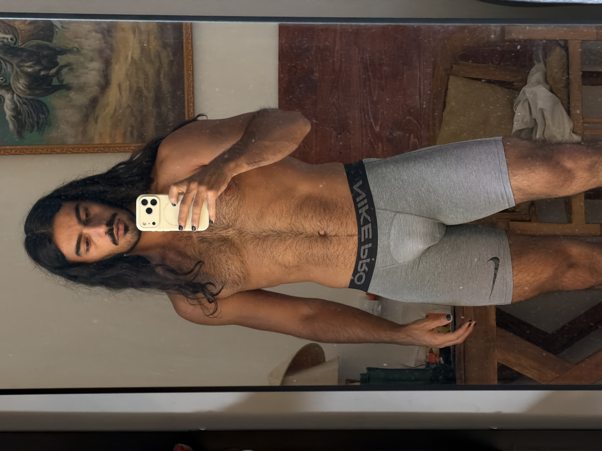 A shirtless man with long, dark, wavy hair and a mustache is taking a mirror selfie in what appears to be a bedroom. He is wearing gray Nike boxer briefs. His left arm is raised, holding a white smartphone. The room has wooden paneling on the wall behind him, and a framed painting of horses hangs on the wall. A lamp with a straw shade is visible to the left, and a wooden chair with a cushion is in the background to the right.