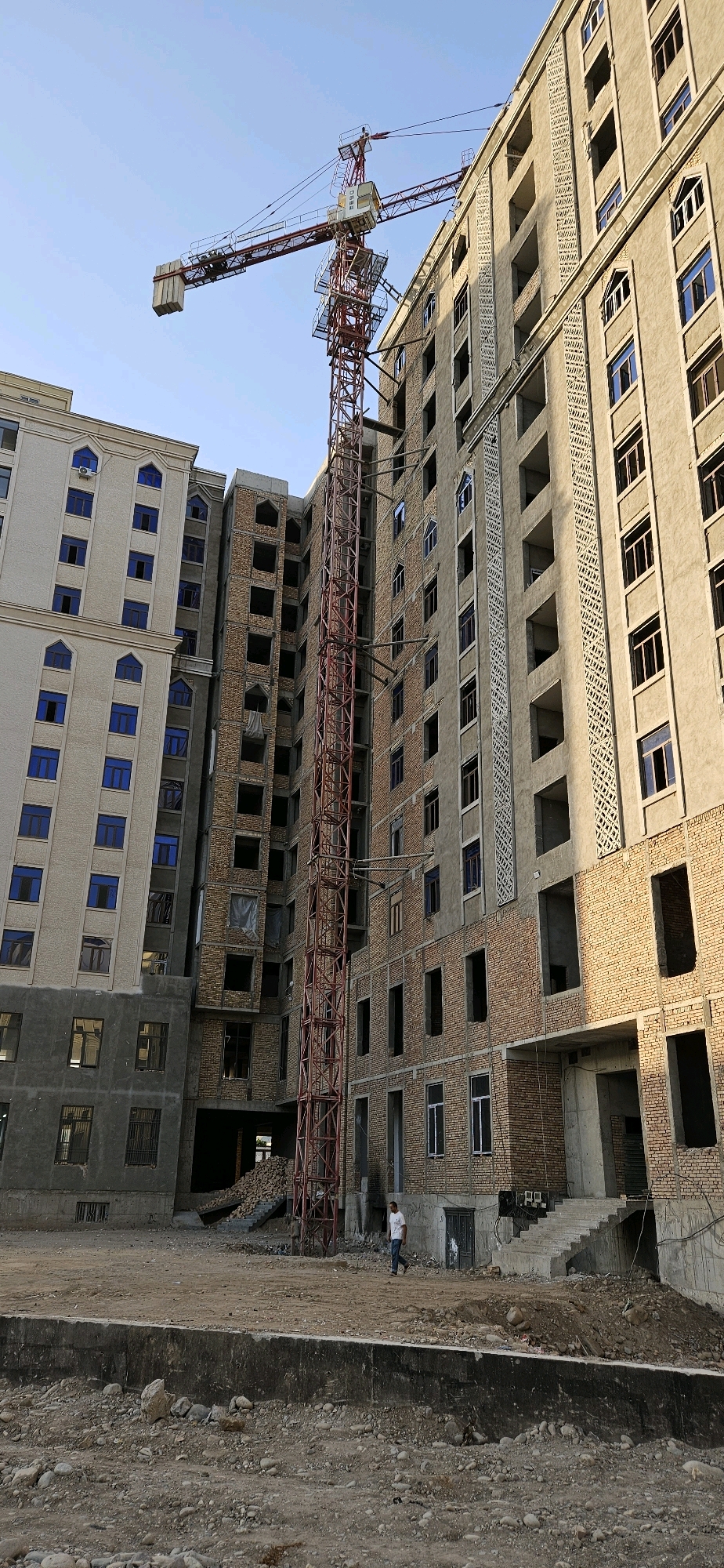 The image depicts a construction site in Dushanbe, Tajikistan, featuring a large tower crane extending over unfinished apartment buildings. The buildings are in various stages of construction, with exposed brickwork and empty window frames visible. One building on the left appears more completed with blue-tinted windows. A single person is walking across the dusty, rocky ground in front of the buildings. The sky is clear and blue, indicating daytime. The scene is raw and industrial, typical of ongoing urban development.