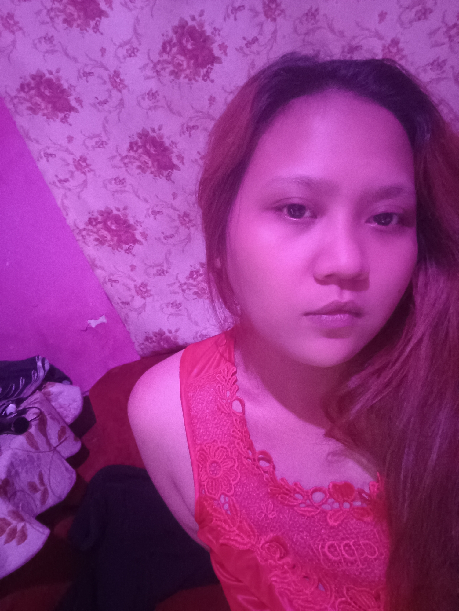 In a bedroom in Depok, Indonesia, a young woman with long, reddish-brown hair is captured in a selfie. She is wearing a bright pink top with intricate lace detailing around the neckline. The background consists of a wall with floral wallpaper in shades of pink and purple. To the left of the woman, a stack of fabric items with a floral pattern is visible. The lighting in the room is predominantly pink, casting a vibrant hue over the scene. The woman's expression is neutral, and her gaze is directed towards the camera.