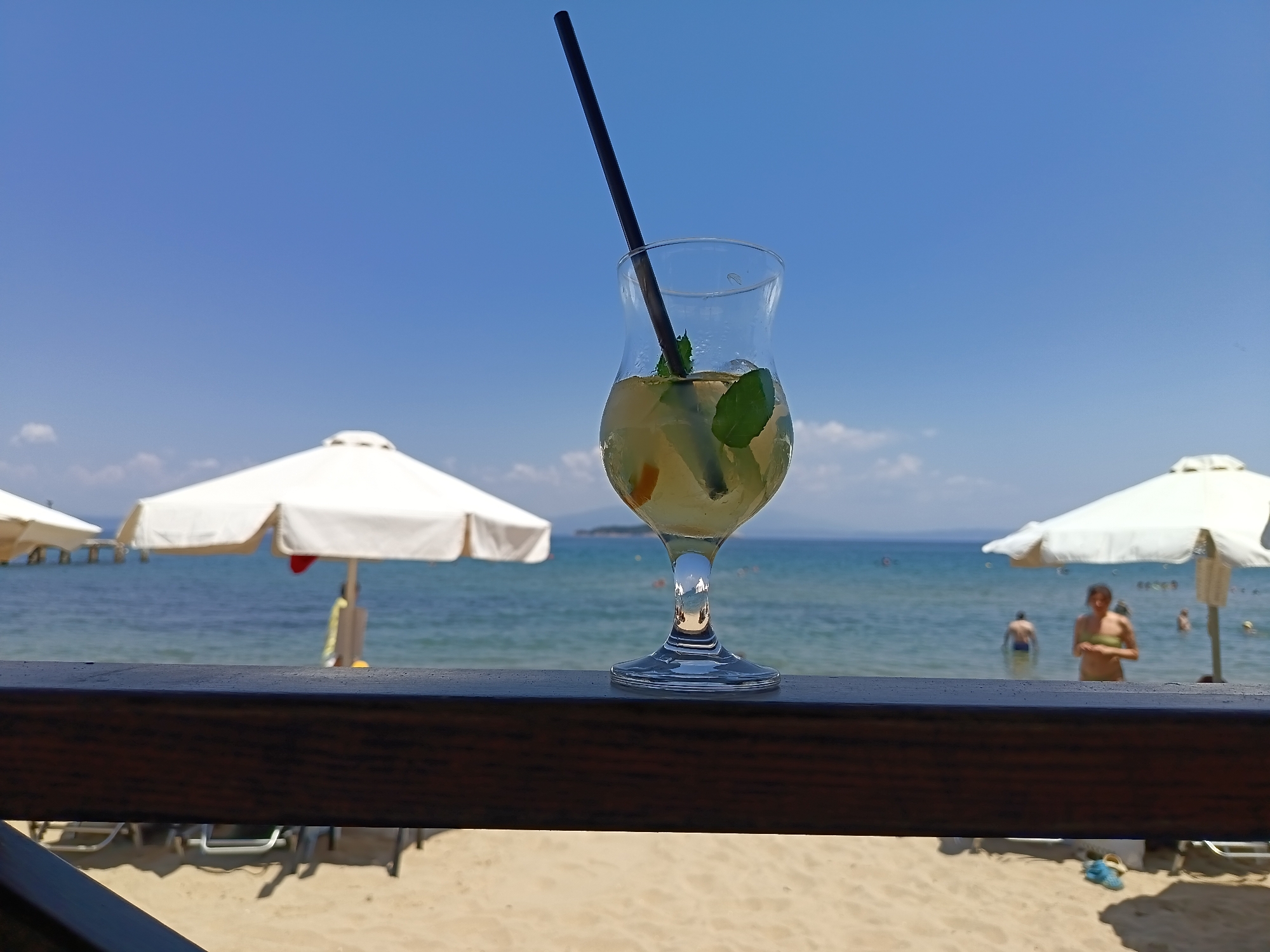 A tall, clear hurricane glass filled with a pale yellow liquid and ice, garnished with mint leaves and citrus slices, stands on a dark wooden railing. A black straw extends from the drink. In the background, the turquoise waters of the Aegean Sea meet a clear blue sky with scattered white clouds. Two white beach umbrellas are visible, one on the left and one on the right, casting shadows on the sandy beach. Several beach chairs are arranged on the sand. In the sea, two men and a woman are visible, with the woman standing and the men wading in the water. The setting appears to be a beachfront cafe or bar in Ολυμπιάδα, Greece, during daylight hours.