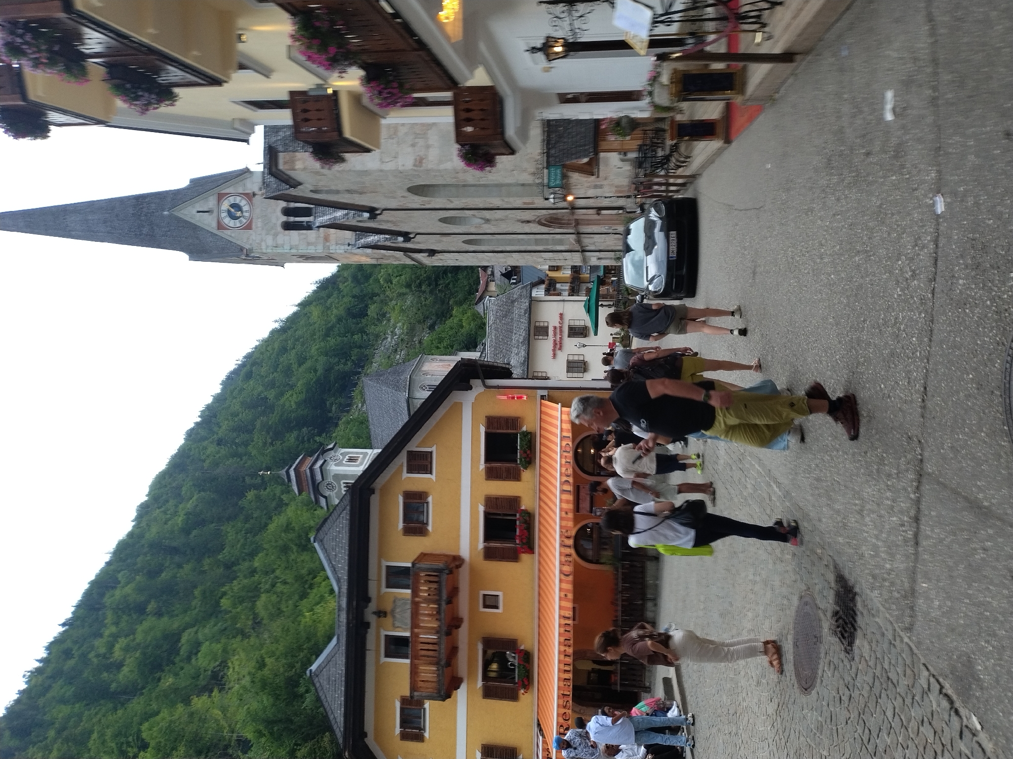The image captures a street scene in the Austrian city of Hallstatt, with a prominent church spire in the background. In the foreground, a cobblestone street is lined with buildings. On the left, a yellow building features balconies and shuttered windows, with an awning covering a "Restaurant Cafe Derbi" entrance. Several people are visible walking and standing on the street. A dark sedan is parked on the right side of the street, in front of a building with outdoor seating. The environment appears to be a European town, with dense green hills rising behind the buildings.