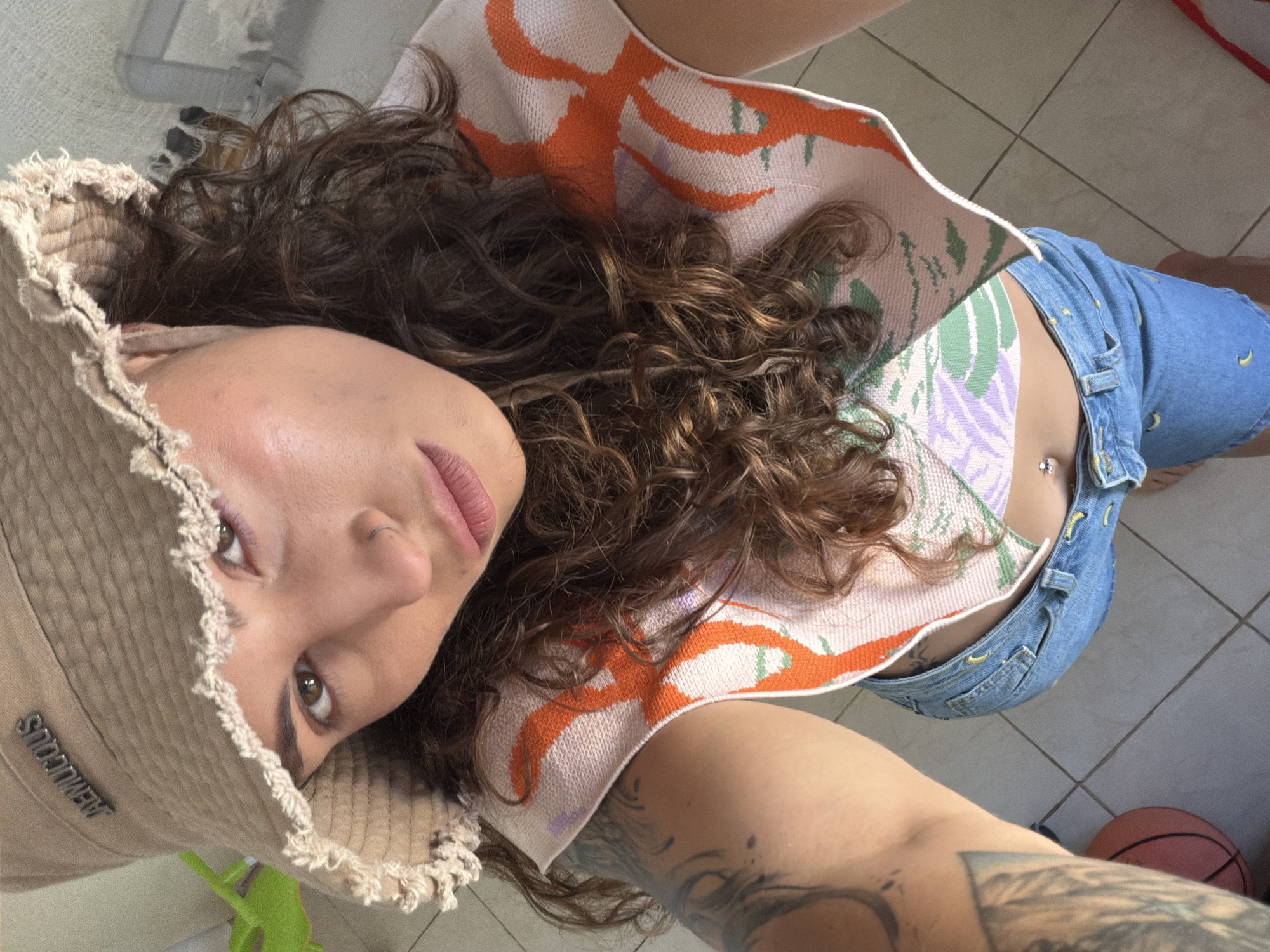 A person is lying down indoors, wearing a beige frayed sunhat with a logo visible on the left side, a patterned cropped shirt in pastel orange, green, and purple, and blue denim shorts with yellow accents. A navel piercing is visible. The person has curly brown hair, a nose ring, and is looking into the camera. Visible tattoos are on the person's left arm. The background includes floor tiles, part of a green object, and a pink basketball. The location is unspecified and there is no information from the image that it is in Kiryat Bialik, Israel.