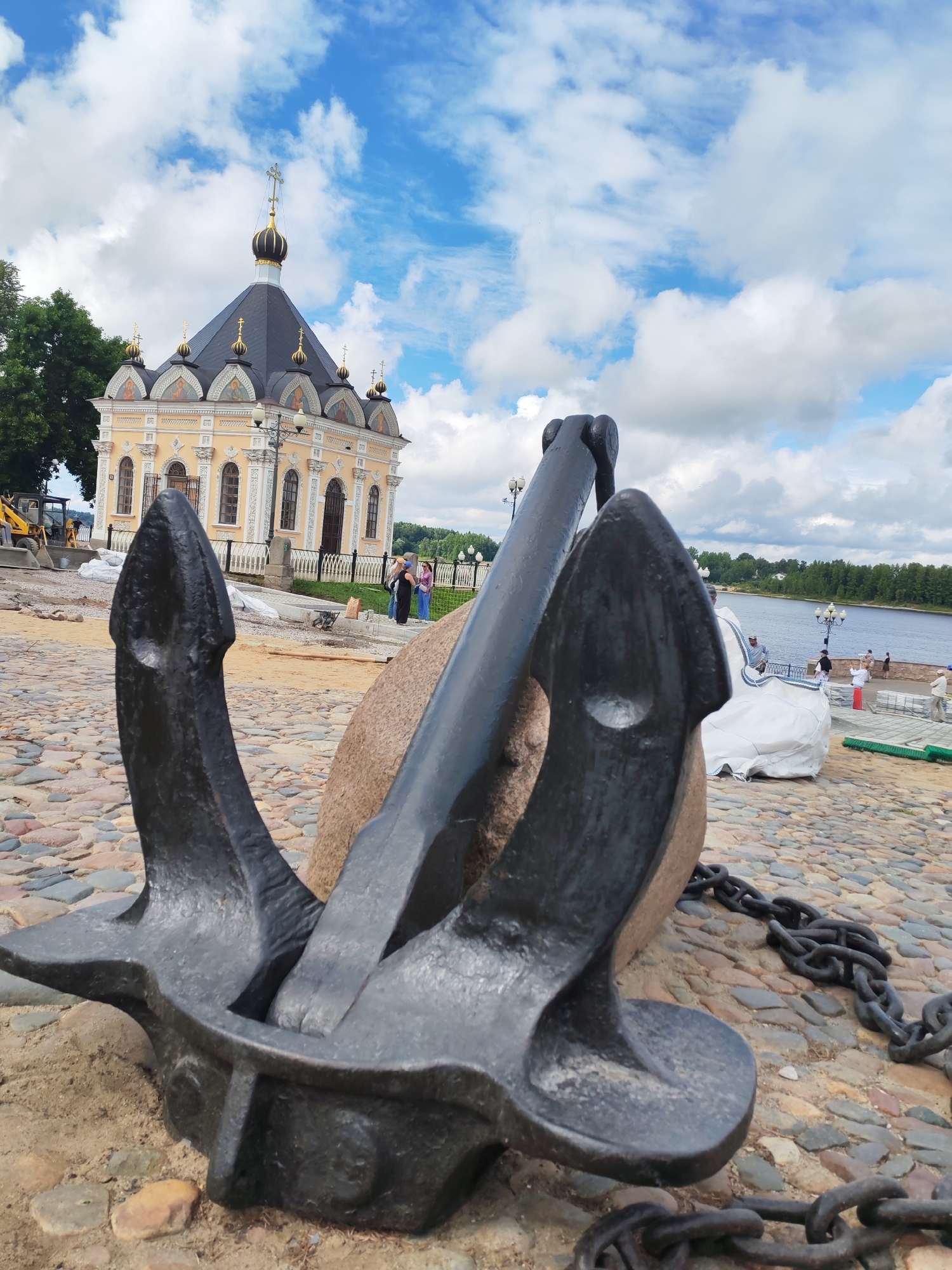 The image depicts an outdoor scene at the city of Bol'shie Mkhi, Russia. In the foreground, a large, dark metal anchor rests on a cobblestone ground, supported by a large stone. A heavy chain is visible extending from the anchor. Behind the anchor, a pale yellow building with a dark roof and golden domes is prominent. The building features arched windows and ornate architectural details. Further in the background, a wide body of water, likely a river, is visible with trees lining the far bank. Several people are present in the scene, some standing near the building and others walking along the waterfront. The sky above is blue with scattered white clouds. Construction materials, such as white sheeting and landscaping elements, are also present on the ground near the building, suggesting ongoing development or renovation.