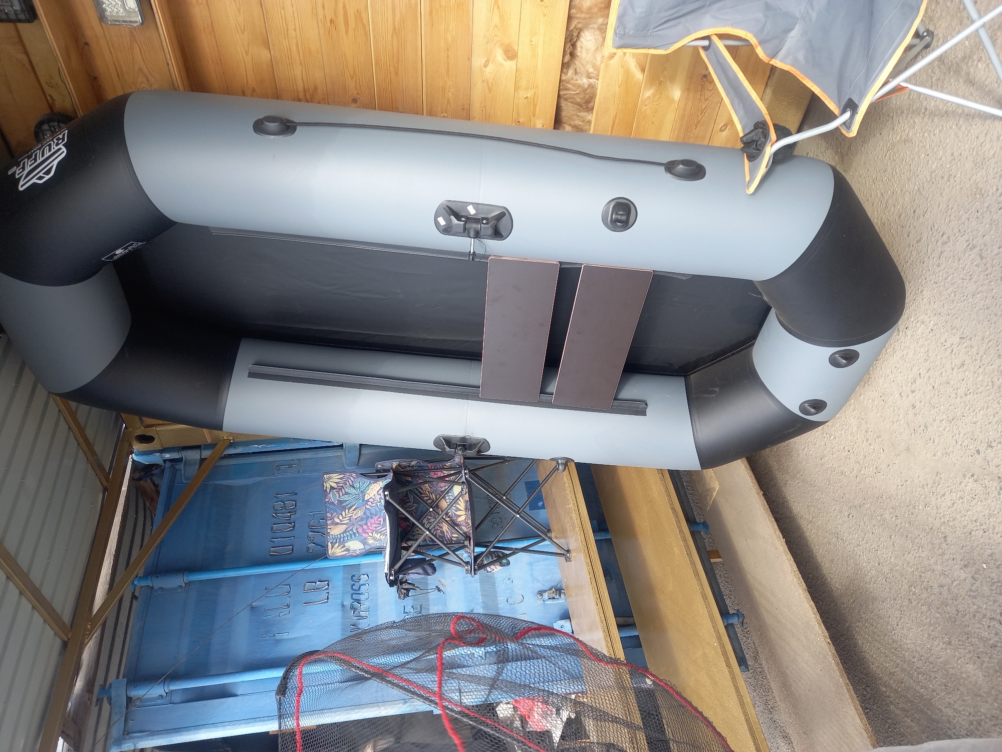 The image shows an inflatable boat, gray and black, with two wooden seat boards placed inside. A folding chair and a fishing net are also visible near the boat. The boat and other objects appear to be located in or near a storage container or shed. The location is unknown.