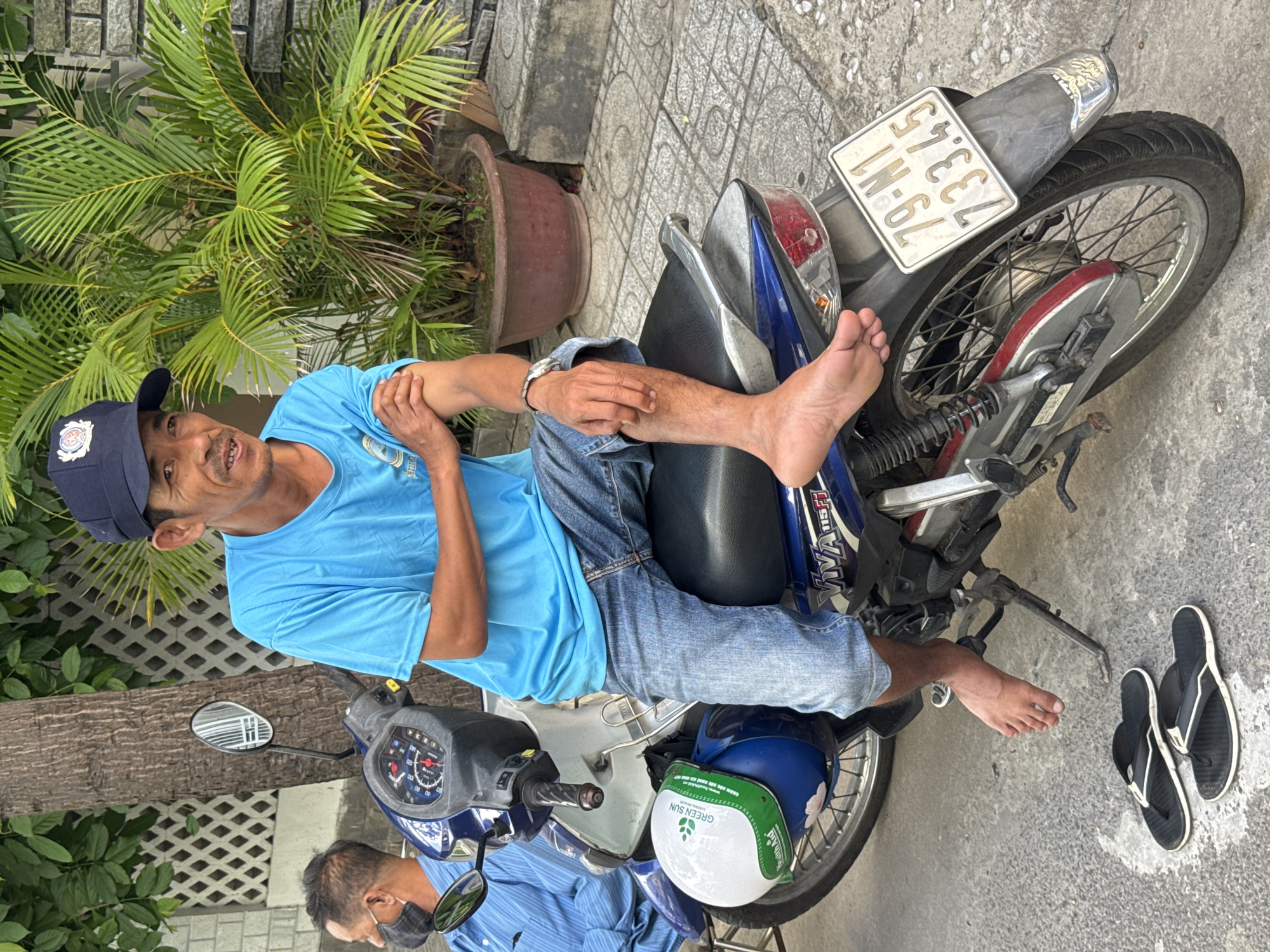 A Vietnamese man in a blue t-shirt and denim shorts sits on a blue and white motorcycle. He wears a dark blue baseball cap and has a watch on his left wrist. His right leg is bare, and his feet are resting on the ground. Next to the motorcycle are a pair of black flip-flops. In the background, a person wearing a blue striped shirt and a face mask is visible. A large potted palm plant and a textured wall complete the scene. The license plate on the motorcycle reads "79-N1 73345". The image appears to have been taken on a street in Nha Trang Ward, Vietnam, as indicated by the license plate format and the general environment.