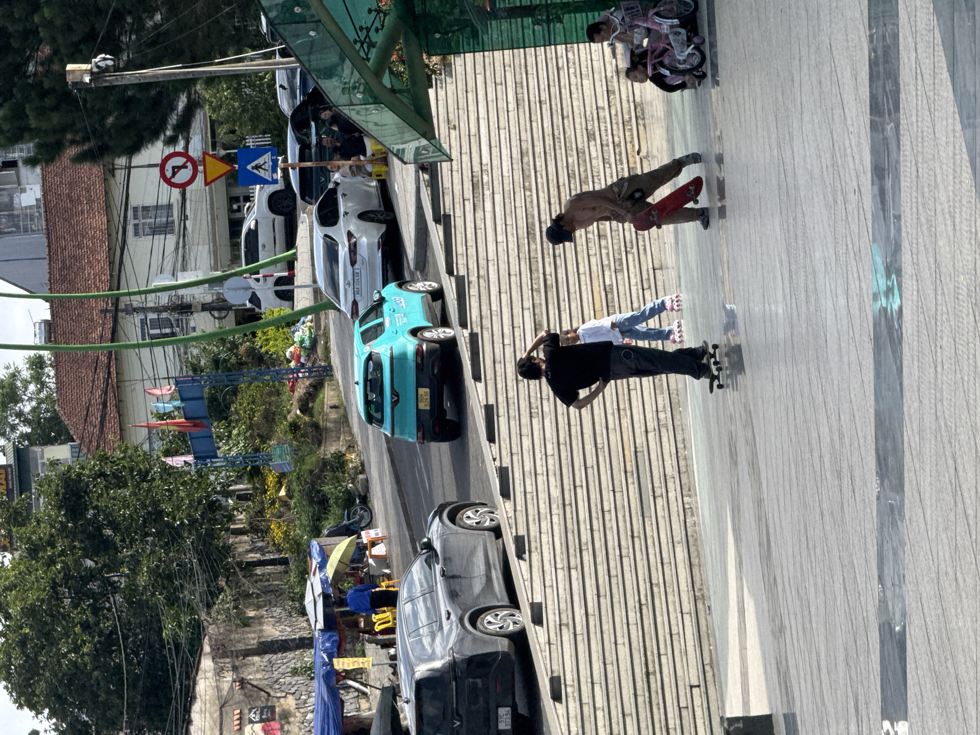 This image depicts a street scene in Da Lat, Vietnam. In the foreground, a man with a shirtless torso and wearing tan pants carries a red skateboard. To his left, a young girl wearing a white shirt and blue pants is on roller skates. A skateboarder on a black skateboard is to the left of the roller-skating girl. In the middle ground, several cars are parked on a street that runs alongside a wide set of concrete stairs. A teal-colored car is parked on the street, and to its left is a dark grey SUV. Further up the street, a white sedan is parked. People are visible sitting on yellow stools under a blue awning. In the background, buildings with red tile roofs are visible, along with trees and street signs. The environment appears to be a sunny day.