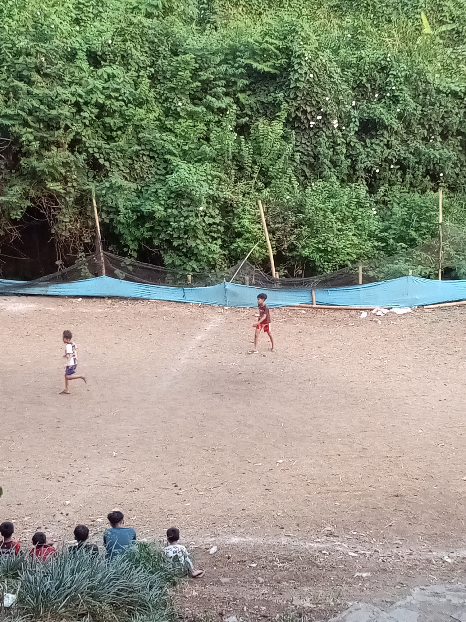 In Sariwangi, Indonesia, a dusty field serves as a makeshift sports area bordered by dense green foliage and bamboo poles supporting a blue tarp and netting. Two young boys are visible on the field, one running to the left in a white and blue shirt and dark shorts, and another standing in the center wearing a red t-shirt and red shorts. In the foreground, a group of children are seated, facing away from the camera, looking towards the field. The ground is uneven and covered with dirt and debris.