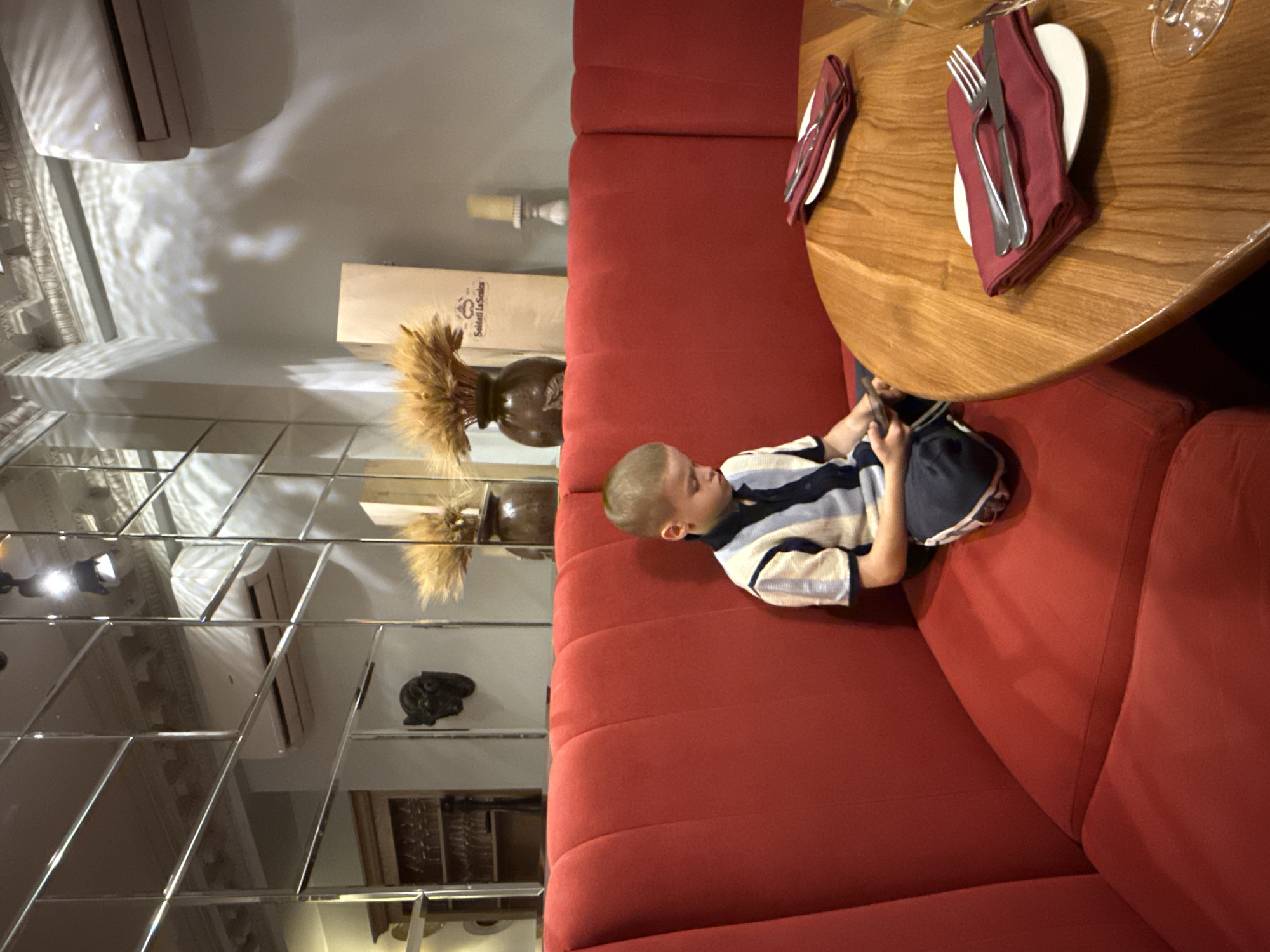 A young boy sits alone on a red upholstered banquette in a dimly lit restaurant. He is looking down at his phone, his legs crossed. The boy is wearing a collared shirt with blue and white stripes and dark pants. To his right is a round wooden table with place settings of folded red napkins and silverware on white plates. The restaurant's interior is reflected in a large mirrored wall behind the banquette. The reflections show a vintage-style air conditioner, decorative moldings on the ceiling, and a wine rack. In the background, there are vases of dried wheat stalks and a paper bag with text that reads "Soldati La Scola." The overall ambiance suggests a sophisticated dining establishment.