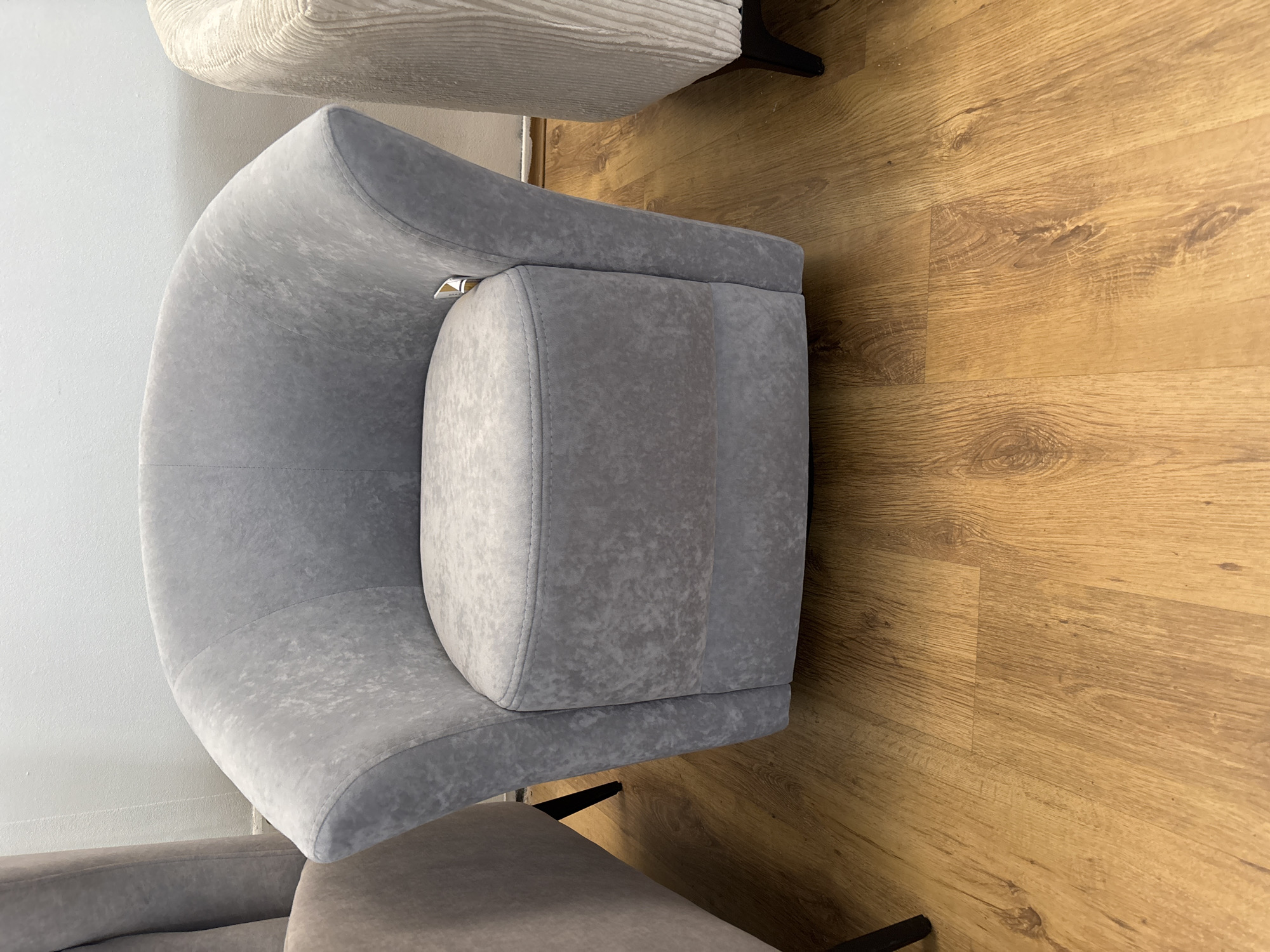 A light gray upholstered armchair with a curved back and seat is centrally positioned on a light brown wooden floor. The armchair has a modern, slightly rounded design. To the left of the main armchair, a portion of a light brown upholstered chair is visible, and to the right, a portion of a cream-colored ribbed upholstered chair is visible. The background consists of a white wall. The image appears to be taken indoors, likely in a furniture showroom. The location in Zaporizhzhia, Ukraine is not determinable from the image content.