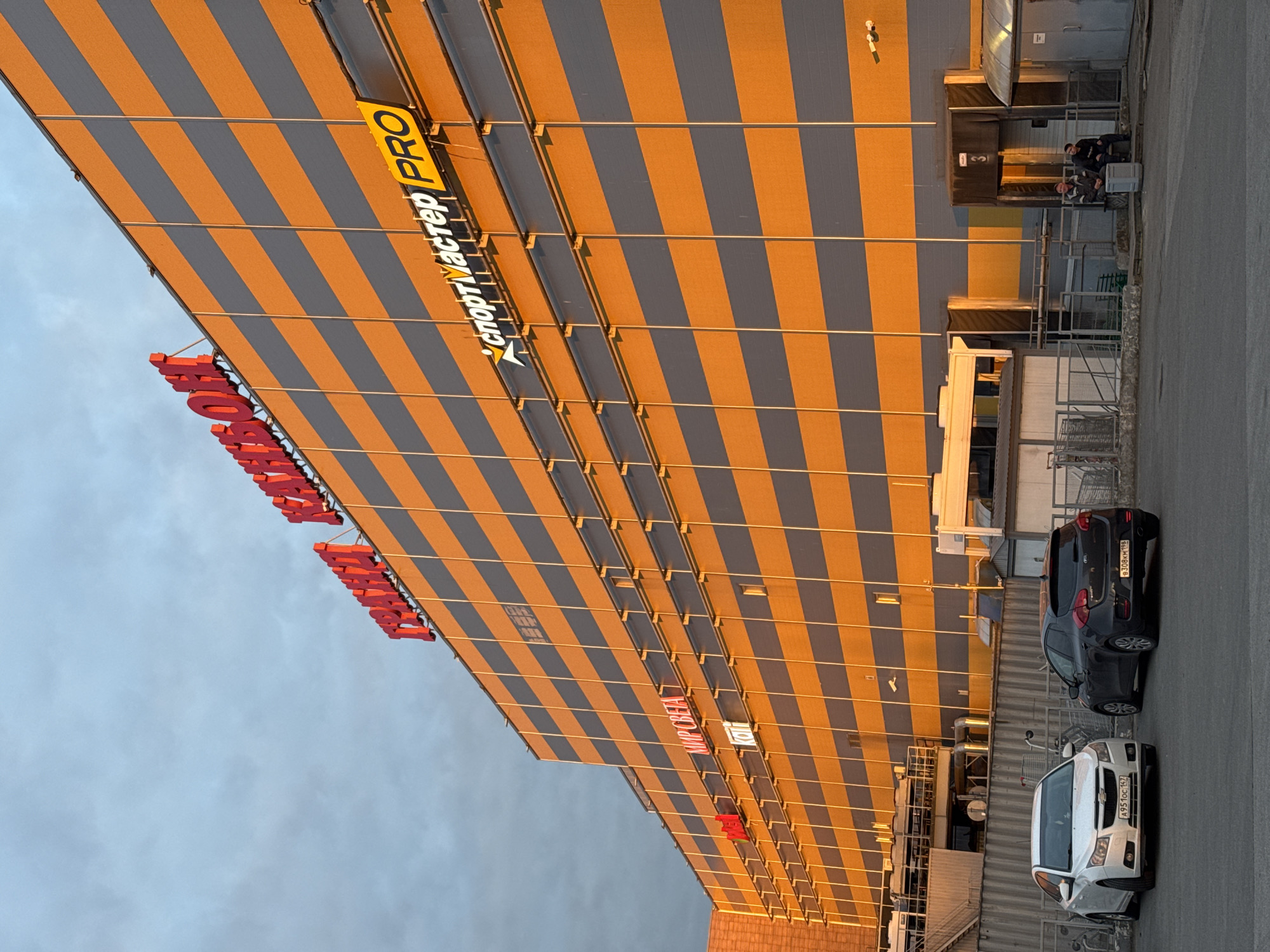 A large commercial building with a distinctive facade featuring alternating stripes of warm orange/gold and dark grey/blue dominates the scene. The image, which appears to be rotated, depicts the building under what looks like a warm sunrise or sunset, casting long, soft light across its surfaces.
Prominently displayed on the building are large signs. A bright yellow and white sign reads "СПОРТМАСТЕР PRO" (SPORTMASTER PRO), with "СПОРТМАСТЕР" in Cyrillic and "PRO" in Latin script. Other red signs, also in Cyrillic, indicate "СПОРТМАСТЕР," suggesting a large sports goods retailer. Further down the building, another sign in red and white reads "МИР СВЕТА" (World of Light), and a partial sign shows "KOIT."
Near what appears to be a service entrance or loading dock (marked with a "3"), three people are visible. One individual sits on a crate or bench, while two others stand nearby, seemingly taking a break or waiting. The area around them includes industrial-looking structures, air conditioning units, and a metal railing.
On the asphalt pavement in front of the building, two cars are parked. The car closer to the foreground is a white Chevrolet, and behind it is a dark-colored hatchback. Both cars bear Russian license plates. Several shopping carts are scattered in the parking area. The sky above is a pale blue with scattered clouds, complementing the warm tones of the building facade.