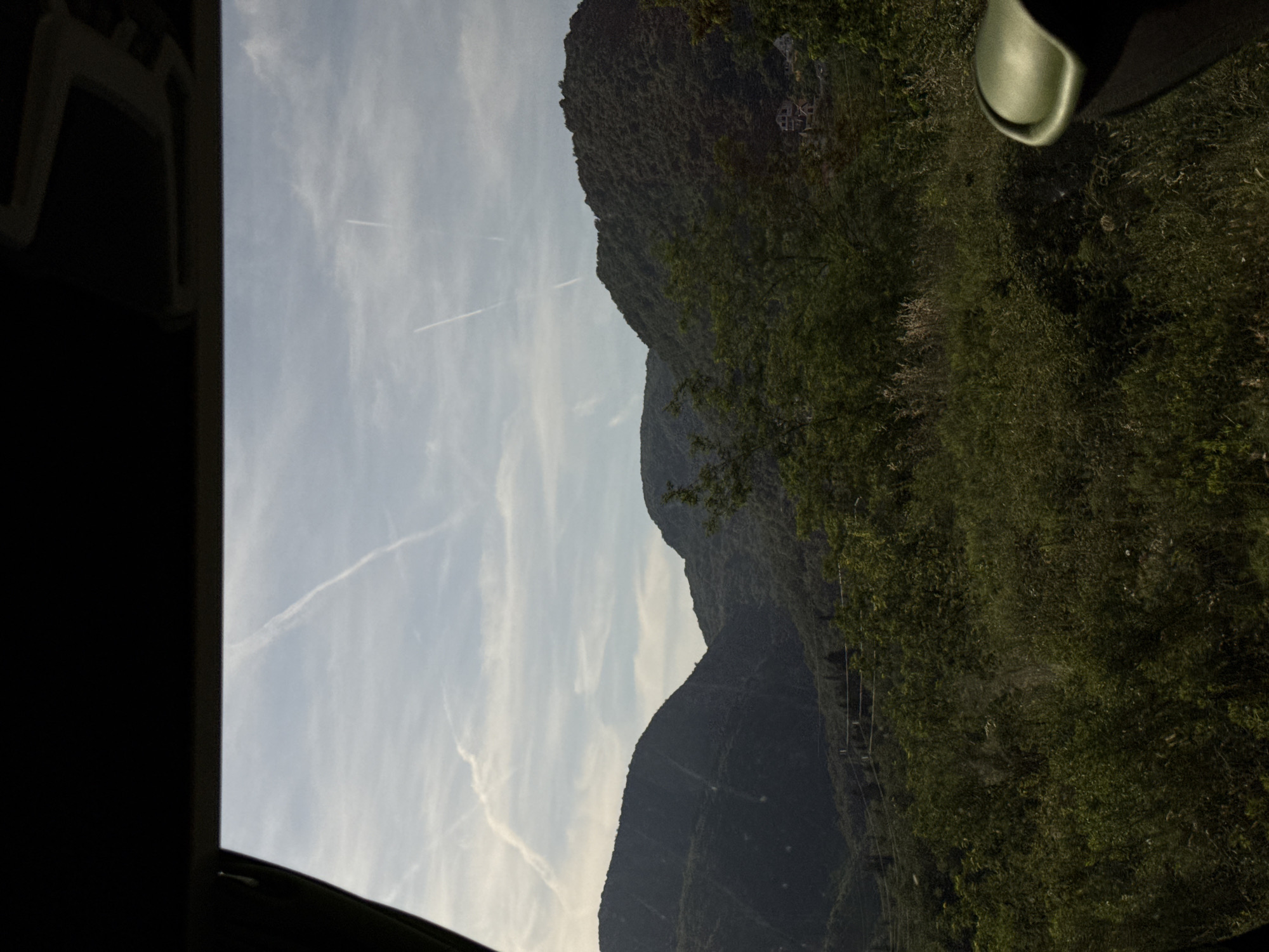 The image shows a view of a mountain landscape in Markovići, Montenegro, taken from inside a vehicle. The mountains are covered in dense green vegetation. The sky is light blue with some contrails visible. The interior of the vehicle is visible in the foreground, including a black window frame and a green object near the top right corner.