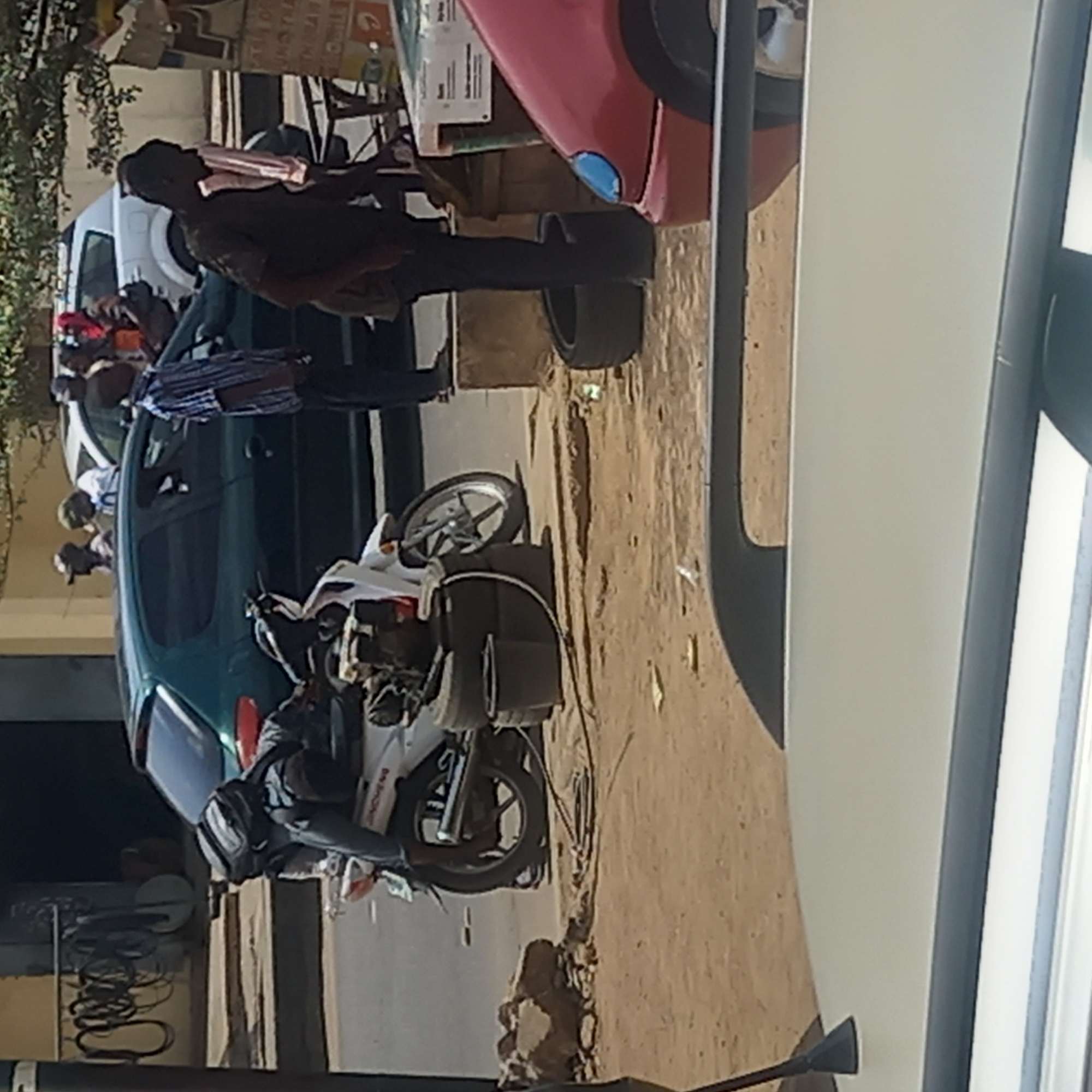 The scene is an outdoor urban setting in Dadi Riba, Nigeria. A green car is parked on a dirt road. A white motorcycle is parked near the car. Several people stand next to the car, including one man leaning against a red car. The environment includes a sandy surface, some vegetation, and buildings.
