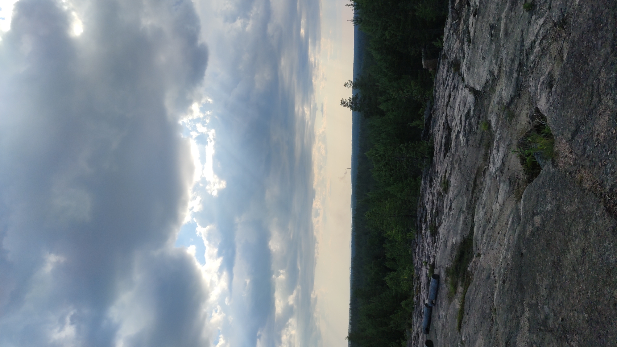 The image depicts a natural landscape with a rocky foreground leading to a dense forest. The sky is filled with dramatic, dark clouds, with patches of bright sky visible, suggesting a transition in weather. In the distance, a thin plume of smoke rises from a chimney, indicating the presence of industrial activity or a dwelling. The overall impression is one of a rugged, natural environment under a dynamic sky. The location is specified as Pitkyaranta, Russia.