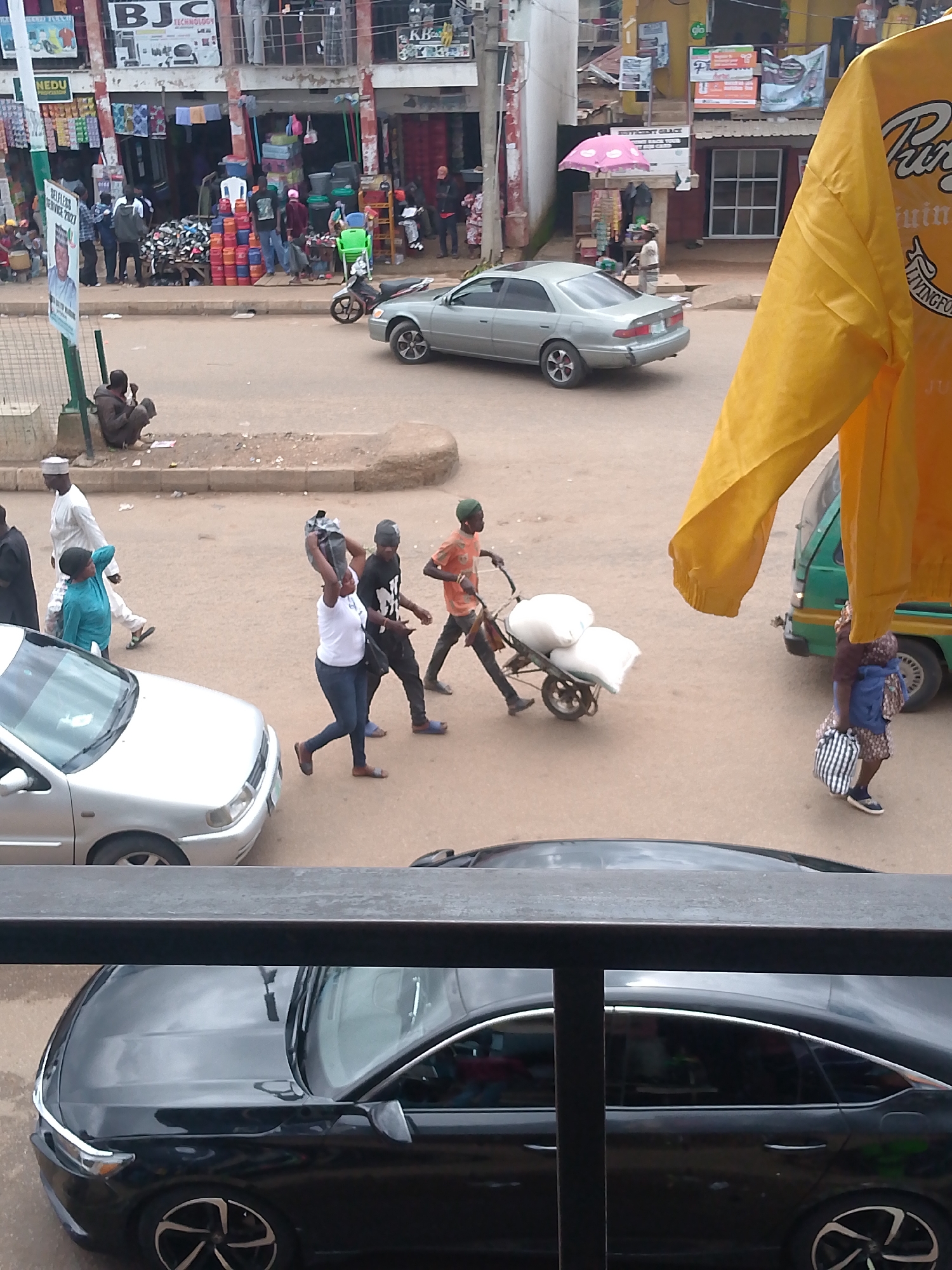 The image depicts a busy street scene in Bukuru, Nigeria, likely in a market area given the presence of shops and vendors. In the foreground, a black car is parked. Above this, a metal railing separates the view from the street. The street itself is populated with various individuals.
Several people are walking on the street. One woman in a white t-shirt and blue jeans is carrying a large black bag on her head and walking with another person. To their right, a man in an orange shirt and green cap is pushing a wheelbarrow laden with two large white sacks. Further back, a silver sedan is driving on the road.
On the left side of the street, a man in a white kaftan and cap is walking. Beside him, a person in a bright blue outfit is also walking. A man dressed in brown is sitting on the edge of the sidewalk. Behind the seated man, a signpost is visible.
The background features a row of shops with awnings and various goods displayed outside. One shop has "BJC TECHNOLOGY" written on its facade. There are also posters and signs attached to the buildings. A pink umbrella is open near one of the shops. A green utility truck is parked on the right side of the street. A person in a patterned dress is walking towards the green truck, carrying a striped bag. A yellow garment is prominently hanging in the upper right portion of the image, partially obscuring the view of the background. The overall environment appears to be an outdoor urban setting with signs of commercial activity and pedestrian traffic.