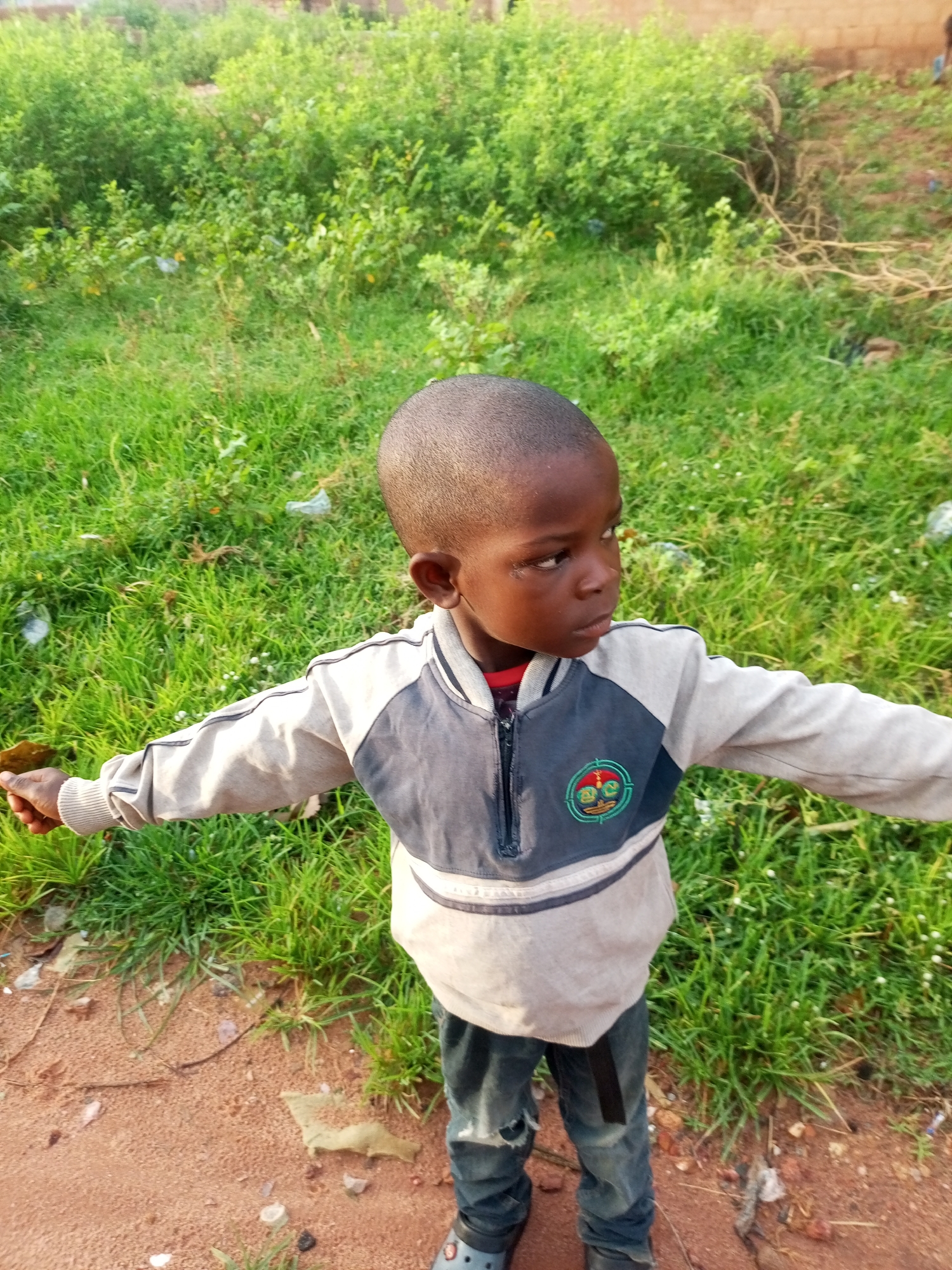 A young boy stands outside in a grassy area, with his arms outstretched. He has a short haircut and is wearing a gray and dark blue jacket over a red shirt, and distressed blue jeans. The background consists of lush green vegetation, and the ground is a mixture of dirt and grass. The scene appears to be in a rural or semi-urban setting in Tempure, Nigeria.