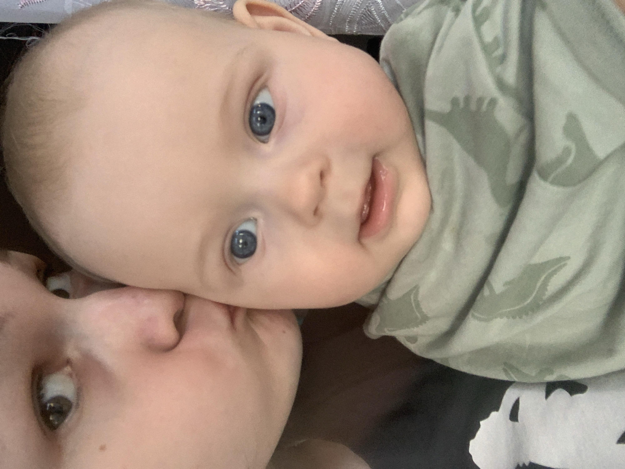 A close-up selfie shows a person kissing a baby on the cheek. The baby has fair skin, blue eyes, and a slightly open mouth. The baby is wearing a light green shirt with a camouflage-like pattern of what appears to be dinosaurs. The person's face is partially obscured by the baby's head. The background is dark and out of focus, with some textured fabric visible on the right. No specific location in Kyiv, Ukraine can be determined from the image.