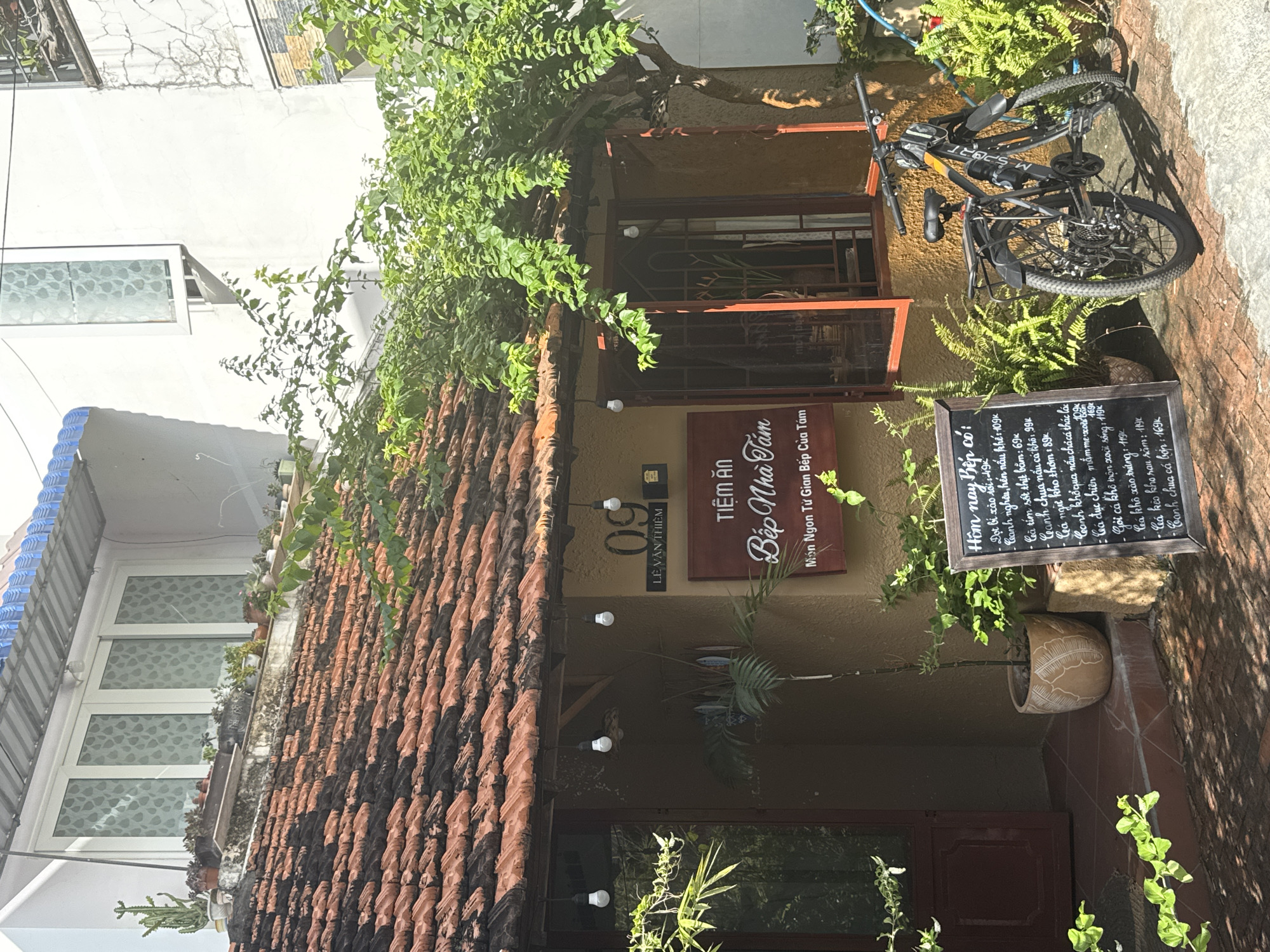 This image depicts the storefront of a Vietnamese restaurant named "Bếp Nhà Tám" (Grandma's Kitchen) located at 09 Lê Văn Thiêm in Phú Thủy, Vietnam. The building features a traditional terracotta tile roof and a muted yellow facade. A large, leafy tree with abundant green foliage partially overhangs the entrance. To the right of the entrance, a bicycle is parked next to a chalkboard menu. The menu lists various dishes with prices in Vietnamese Dong, suggesting a casual dining establishment. The surrounding environment is lush with potted plants and ferns, contributing to a tropical, homey atmosphere. The lighting suggests it is daytime, with natural sunlight casting shadows on the brick-paved ground. The overall scene conveys a sense of a charming, established local eatery in a Southeast Asian city.