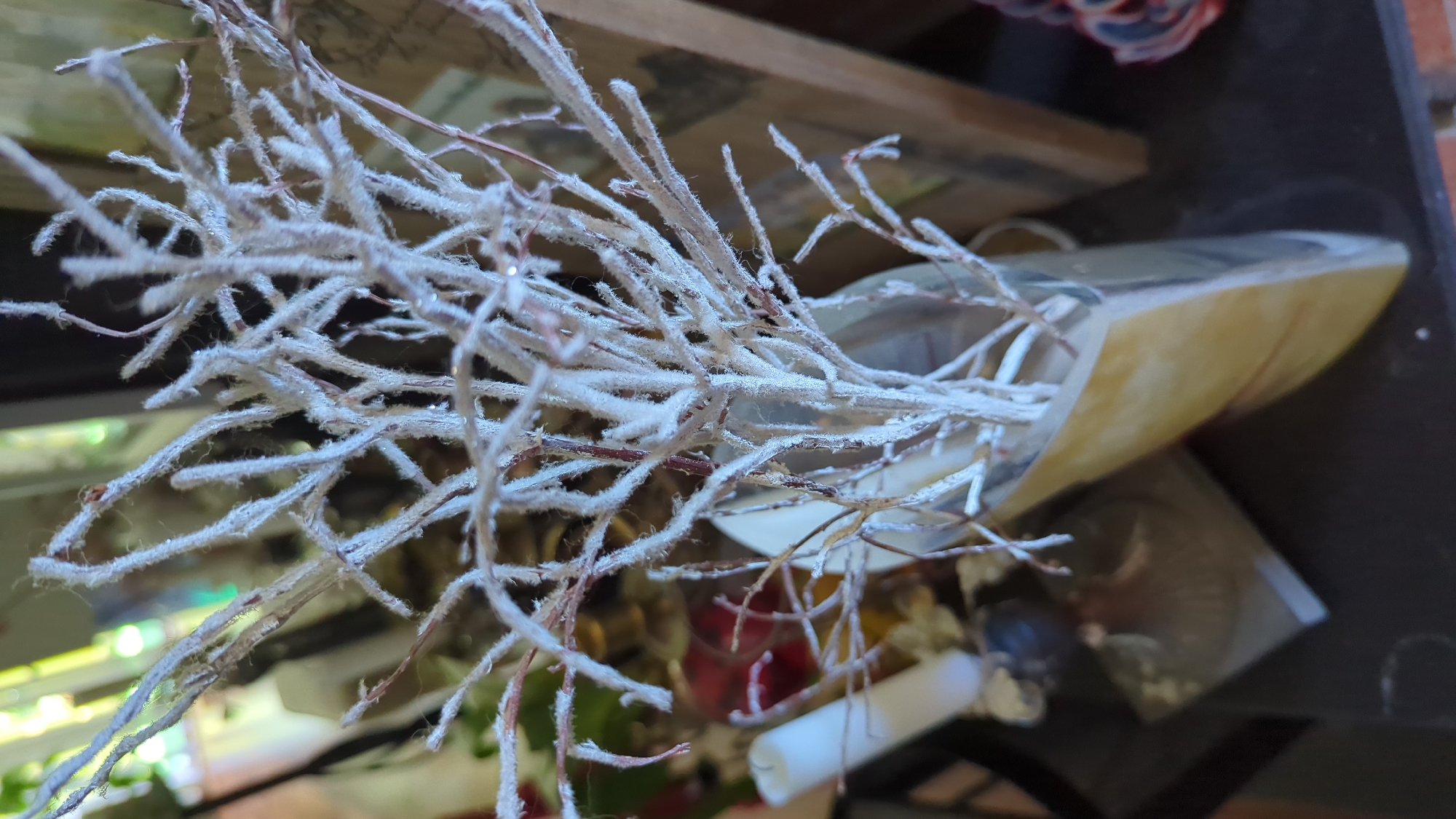This image depicts a close-up view of decorative white-flocked branches in a vase. The branches are intricate and delicate, with a fluffy texture, and some appear to have small water droplets on them. The vase is tall and appears to be made of a material with a yellowish-tan marbling effect, and it has a somewhat angular, modern shape. In the background, out of focus, are hints of other objects, including what looks like a white candle to the left and a wooden frame to the right. The overall setting suggests an indoor environment, possibly a home or a decorative display. The image does not provide any information or context that would allow for its location to be identified as Kløfta, Norway.