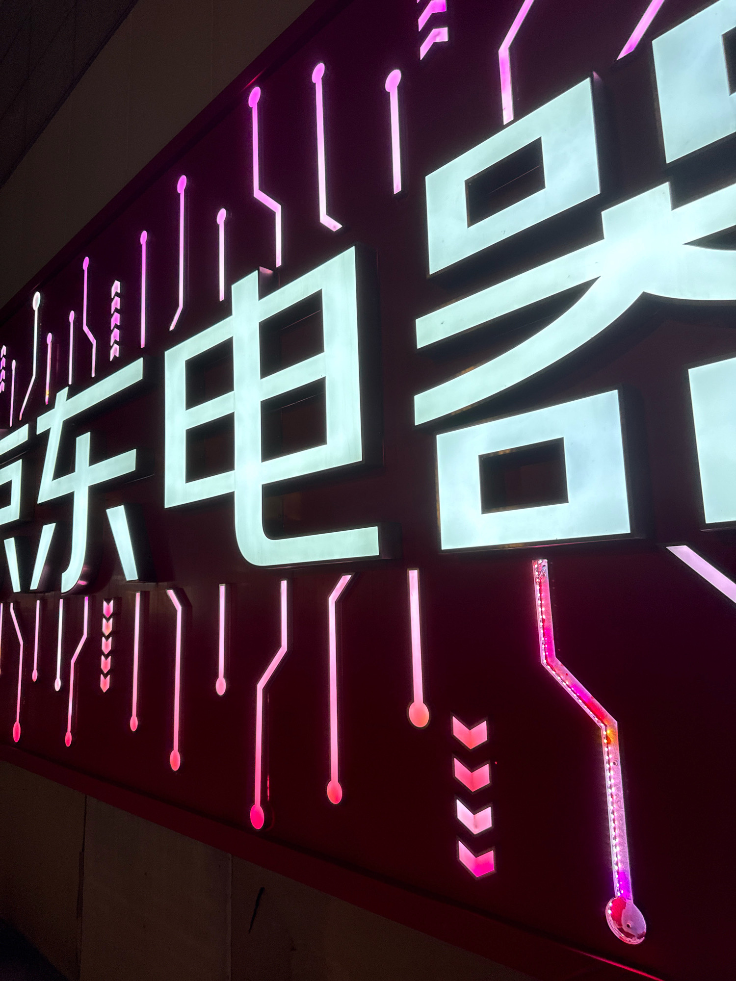 A close-up, angled, eye-level shot captures a neon sign with stylized Chinese characters and circuit board-like accents. The sign is predominantly dark red with bright, illuminated white and pink elements. The main characters, in a bold, blocky font and illuminated in white with a subtle green tint, appear to read "东电器" (Dong Dian Qi), which translates to "East Electric Appliances". These characters are large and three-dimensional, casting faint shadows.
The background of the sign is a deep red. Interspersed with the characters and extending above and below them are thin, vertical pink neon lines with rounded ends, resembling electrical connections or pathways. Some of these lines are straight, while others are angled, creating a visual effect reminiscent of a circuit board. In the lower right portion of the image, a more complex pink neon element snakes diagonally, featuring what appears to be a series of embedded LEDs that transition in color from pink to yellow. Small, chevron-shaped pink arrows are also visible on the dark red surface, arranged in a vertical line. The lighting is artificial and vibrant, creating a high-contrast scene. The top left of the image shows a dark, out-of-focus area, possibly the ceiling or an adjacent structure. No people are visible in the frame.