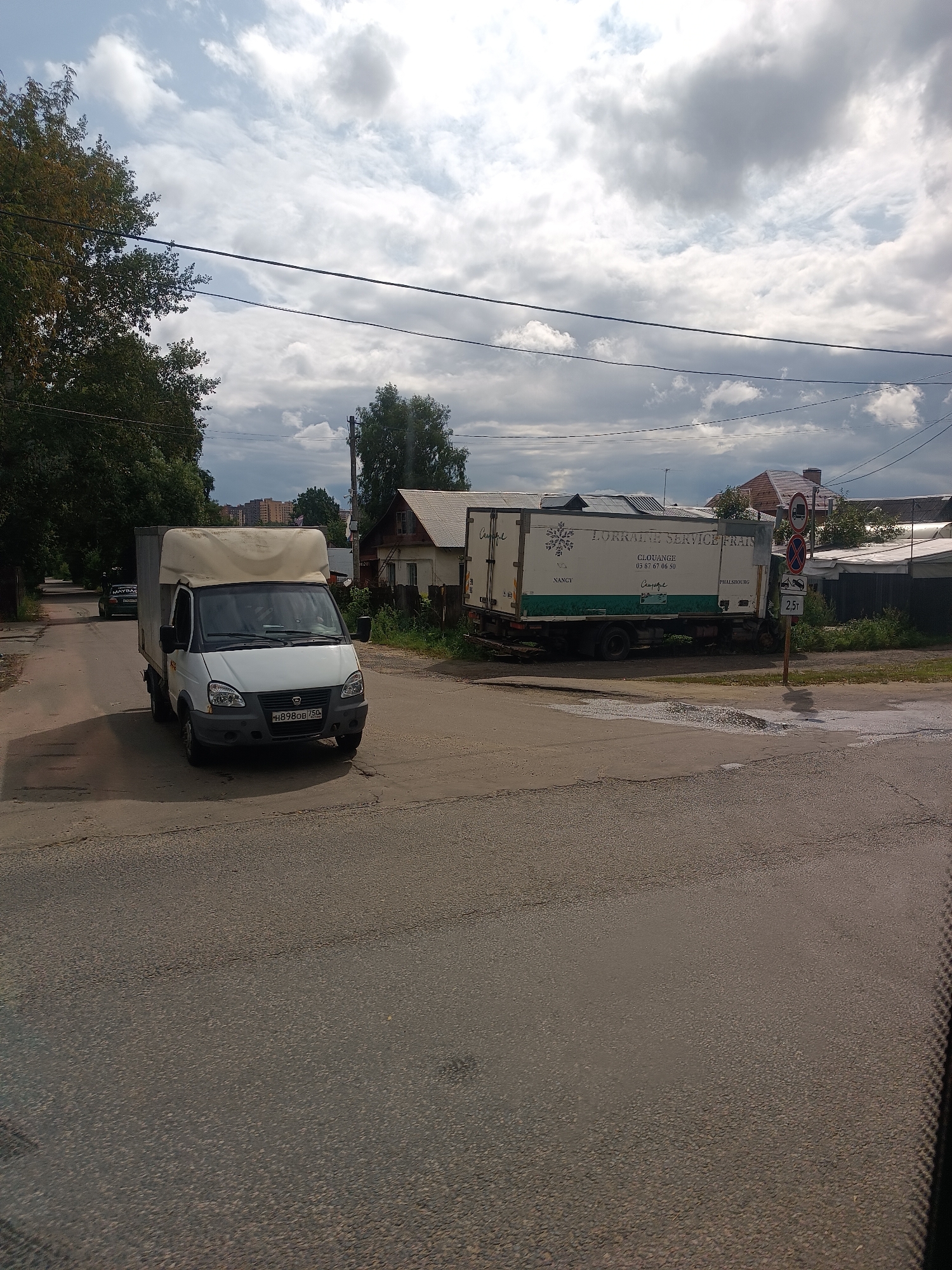 The image depicts a street scene in Chkalovo, Russia, under a cloudy sky. A white cargo van, with a beige tarpaulin cover over its cargo area, is positioned in the foreground, driving on an asphalt road. The van has a license plate that reads "H898OB 750". To the right of the road, there's a light-colored truck with text on its side that includes "LORRAINE SERVICE FRAIS". A traffic sign with multiple indications, including a red circle with a horizontal bar and a red cross, is visible near the truck. Several buildings, including houses with pitched roofs and a taller apartment building in the distance, are present along the street. Trees and power lines are also visible in the environment.