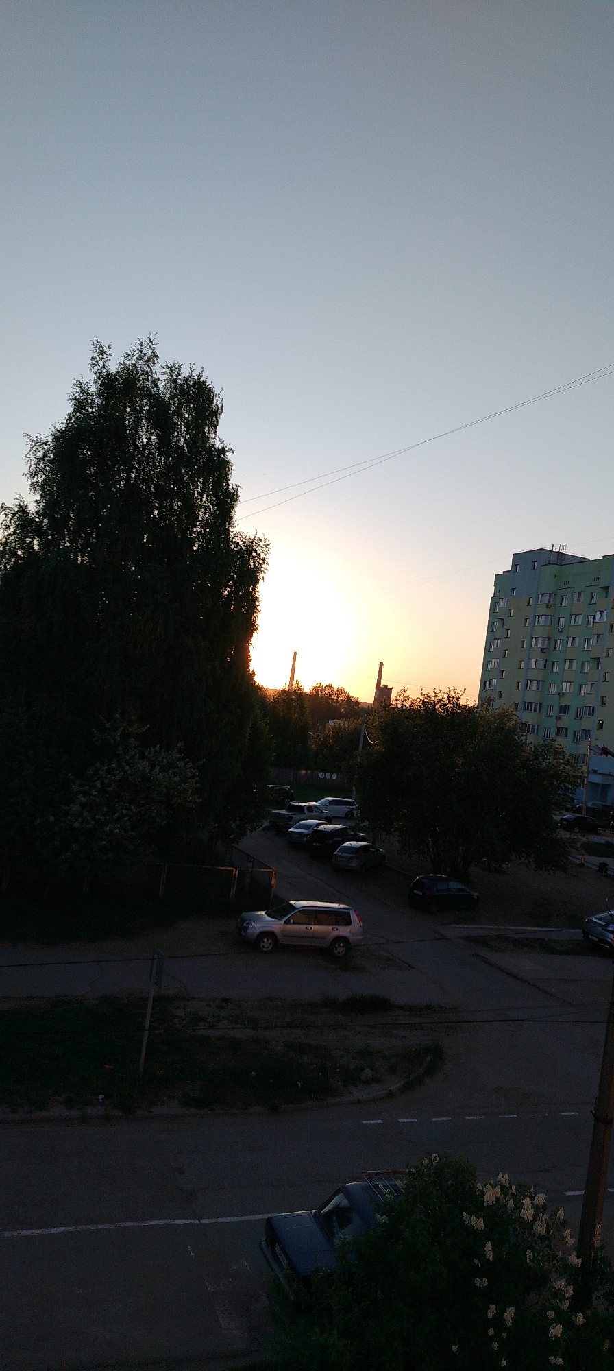 The image captures a sunset in what appears to be a residential area of Chaikovsky, Russia. The sky is a gradient of light blue to orange, indicative of a setting sun. Silhouetted against the sun are two industrial smokestacks. To the left, a tall, dark tree obscures part of the scene. A parking lot is visible with several parked cars of various makes and models. A silver Nissan X-Trail SUV stands out in the foreground. An apartment building with multiple windows rises on the right side. In the bottom right corner, a utility pole and a dark car parked on a street, partially obscured by a flowering bush, are visible. The overall scene is one of a typical evening in a Russian city.