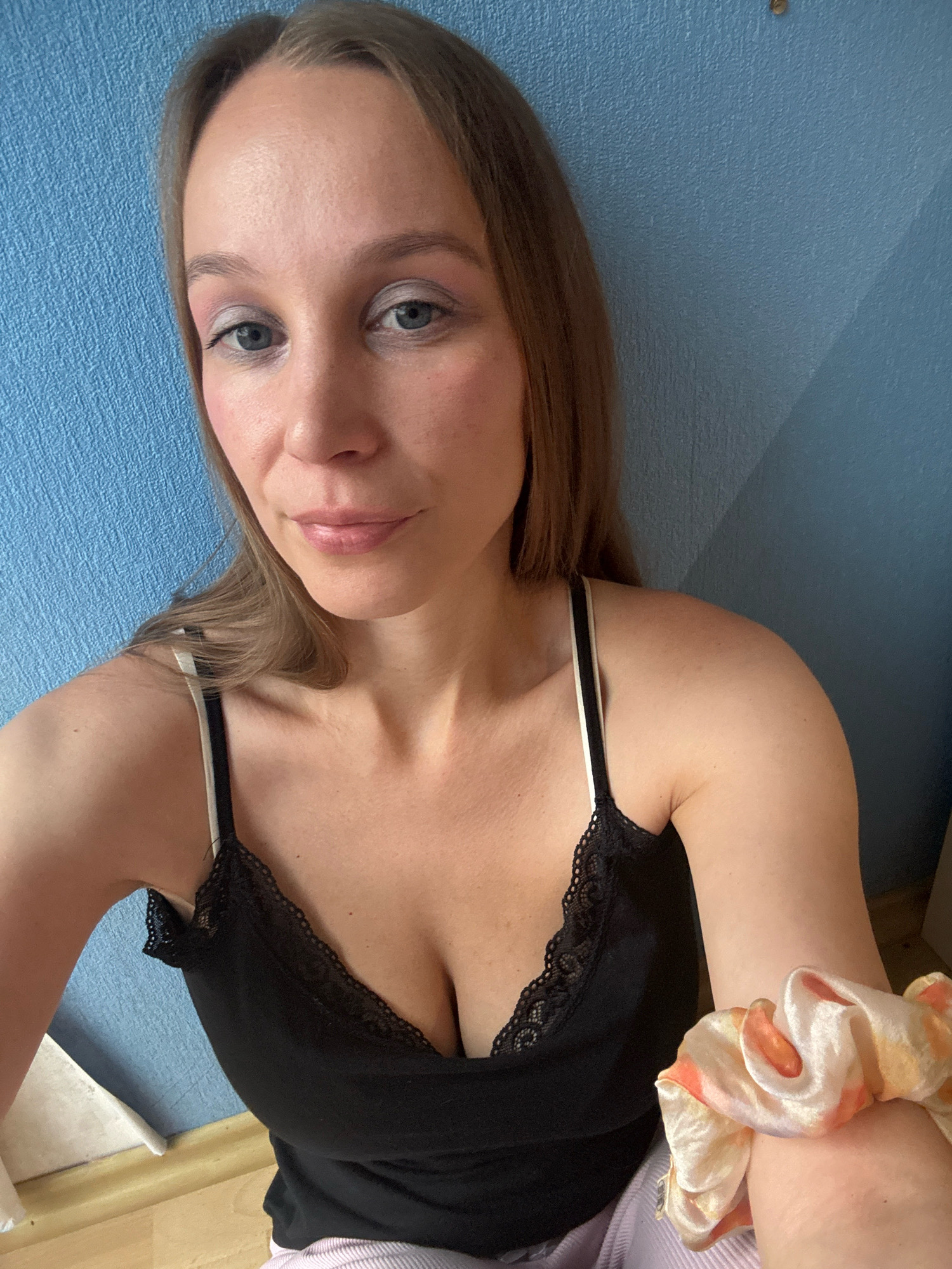 A young woman is taking a selfie against a blue textured wall. She has blonde hair, blue eyes, and is wearing a black tank top with lace trim and light pink pants. She is also wearing a scrunchie on her right arm. The background appears to be indoors, possibly a bedroom or living area, with a floor visible at the bottom of the image. There is no discernible information about the location in the city of Überherrn, Germany.