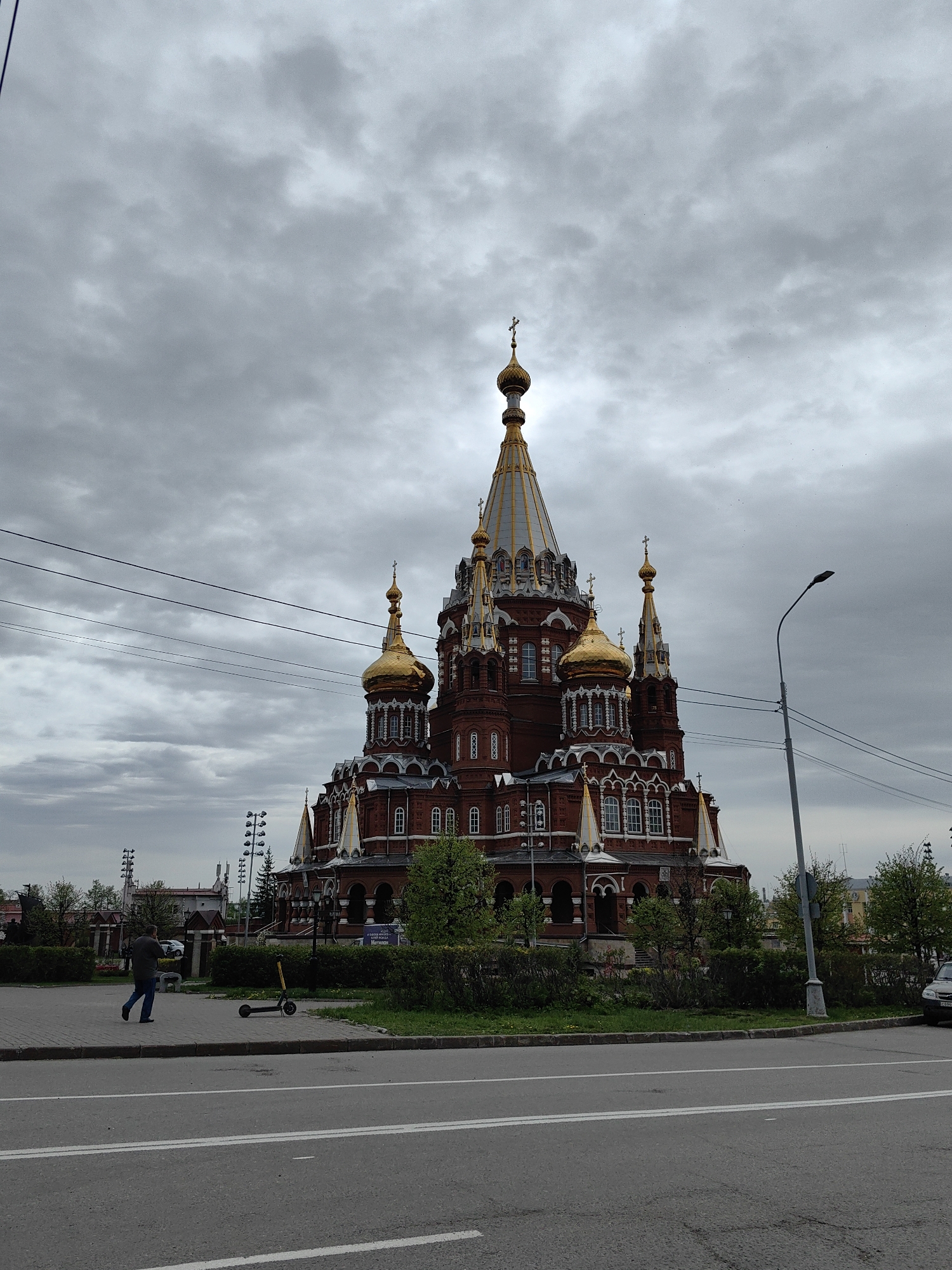 The image shows the Alexander Nevsky Cathedral in Izhevsk, Russia, under a cloudy sky. The cathedral is constructed of red brick with white trim and features multiple golden domes and spires topped with crosses. A lamppost is visible on the right side of the frame. In the foreground, there is a road with white lane markings. A person is walking on the sidewalk next to the road. A parked electric scooter is also visible on the sidewalk. Some greenery (bushes, trees) separates the sidewalk and the cathedral. A car is parked to the right behind the bushes.