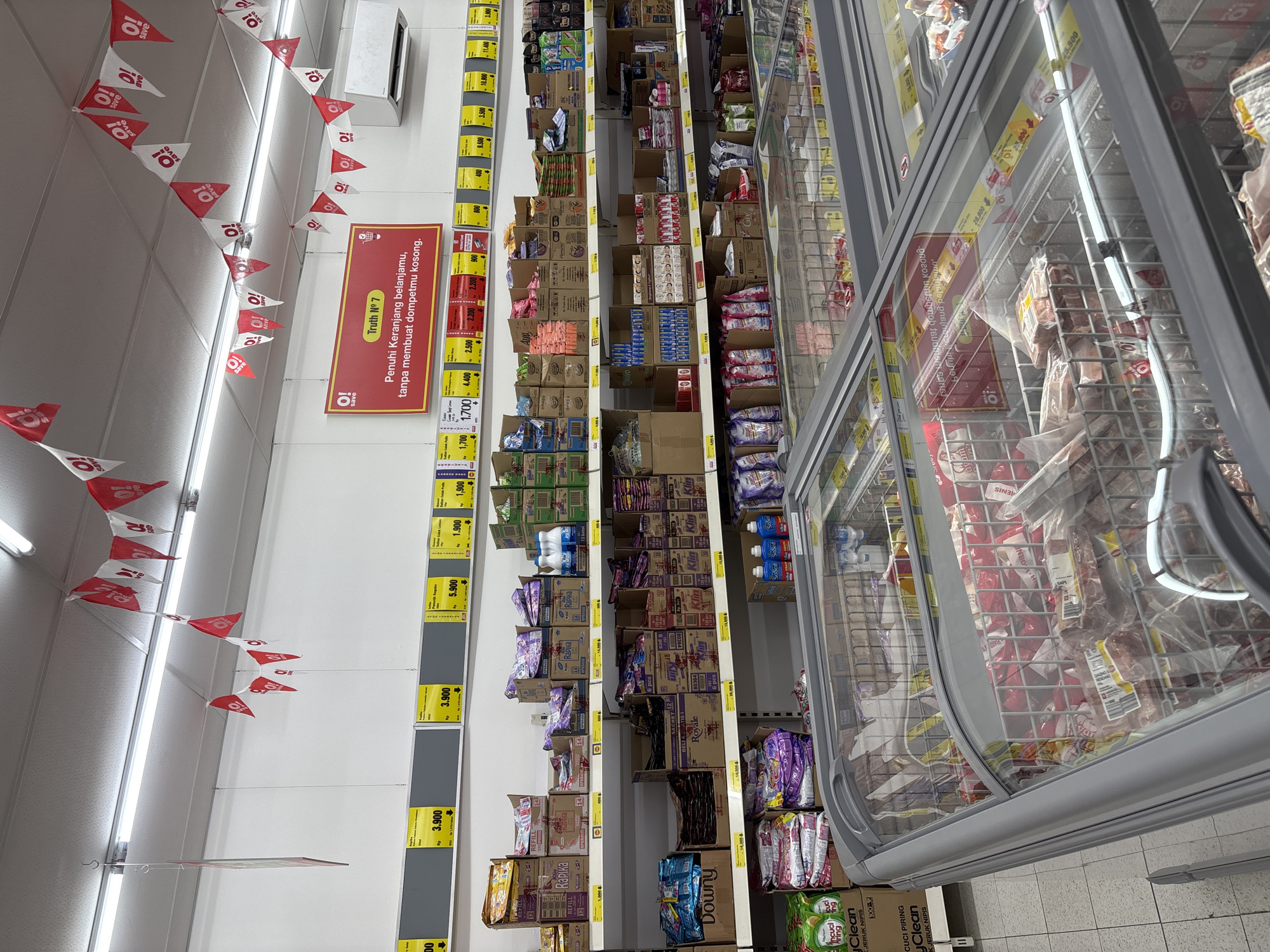 The image depicts a supermarket aisle, likely in Sukabakti, Indonesia, as indicated by the text on the promotional banner. The aisle is stocked with various products, including boxed goods and items inside a refrigerated display case. Red and white triangular flags with the text "O! save" are strung along the ceiling. Yellow price tags are attached to the shelves. A sign in Indonesian reads "Truth No. 7: Fill your shopping basket, without emptying your wallet." The products in the refrigerated case appear to be meat or frozen food. Laundry detergent products are visible on the lower shelves.