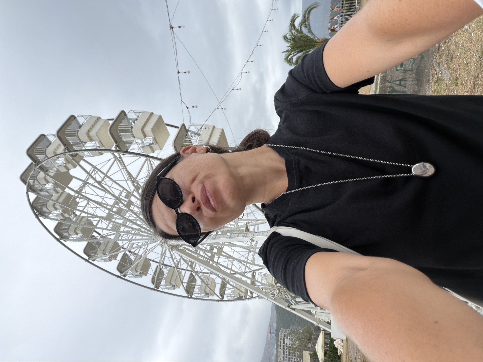 A woman wearing sunglasses, a black t-shirt, and a silver pendant is taking a selfie with a Ferris wheel in the background. The Ferris wheel is white and large, with multiple cabins. String lights are visible above the Ferris wheel, and a palm tree is to the right. In the background, there are some buildings under construction and a cloudy sky. The location is the city of Budva, Montenegro, likely near the coast, given the presence of a palm tree and the coastal landscape.