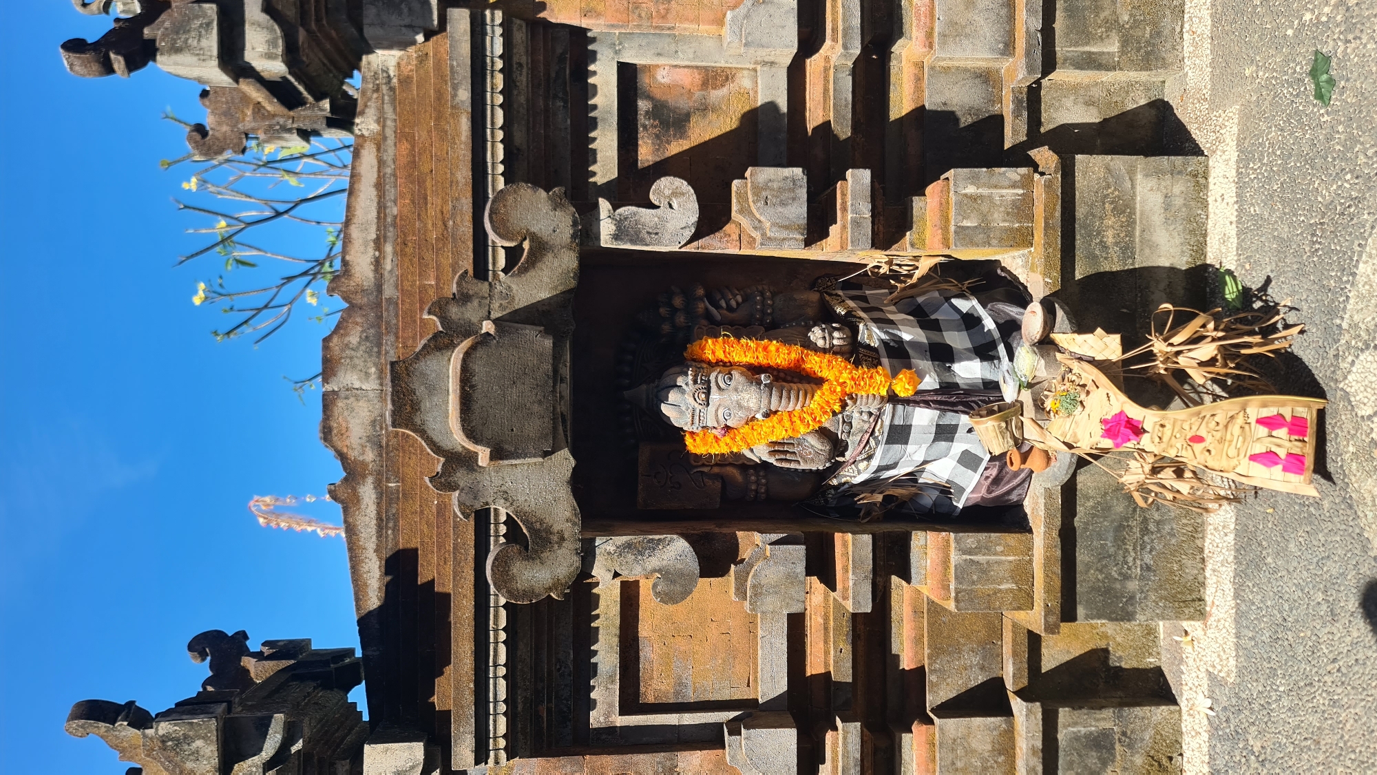 The image shows a stone statue of Ganesha, a Hindu deity, adorned with an orange flower garland and a black and white checkered cloth around its waist. The statue is housed in a small, ornate alcove within a stone structure, likely a temple in Denpasar, Indonesia. The architecture features intricate carvings and is made of weathered stone and bricks. In front of the statue, there is a traditional Balinese offering (Canang Sari) placed on a wooden stand, consisting of dried palm leaves, flowers, and small clay pots. The sky above is a clear, bright blue, and sunlight casts shadows on the stone facade, indicating it is daytime.