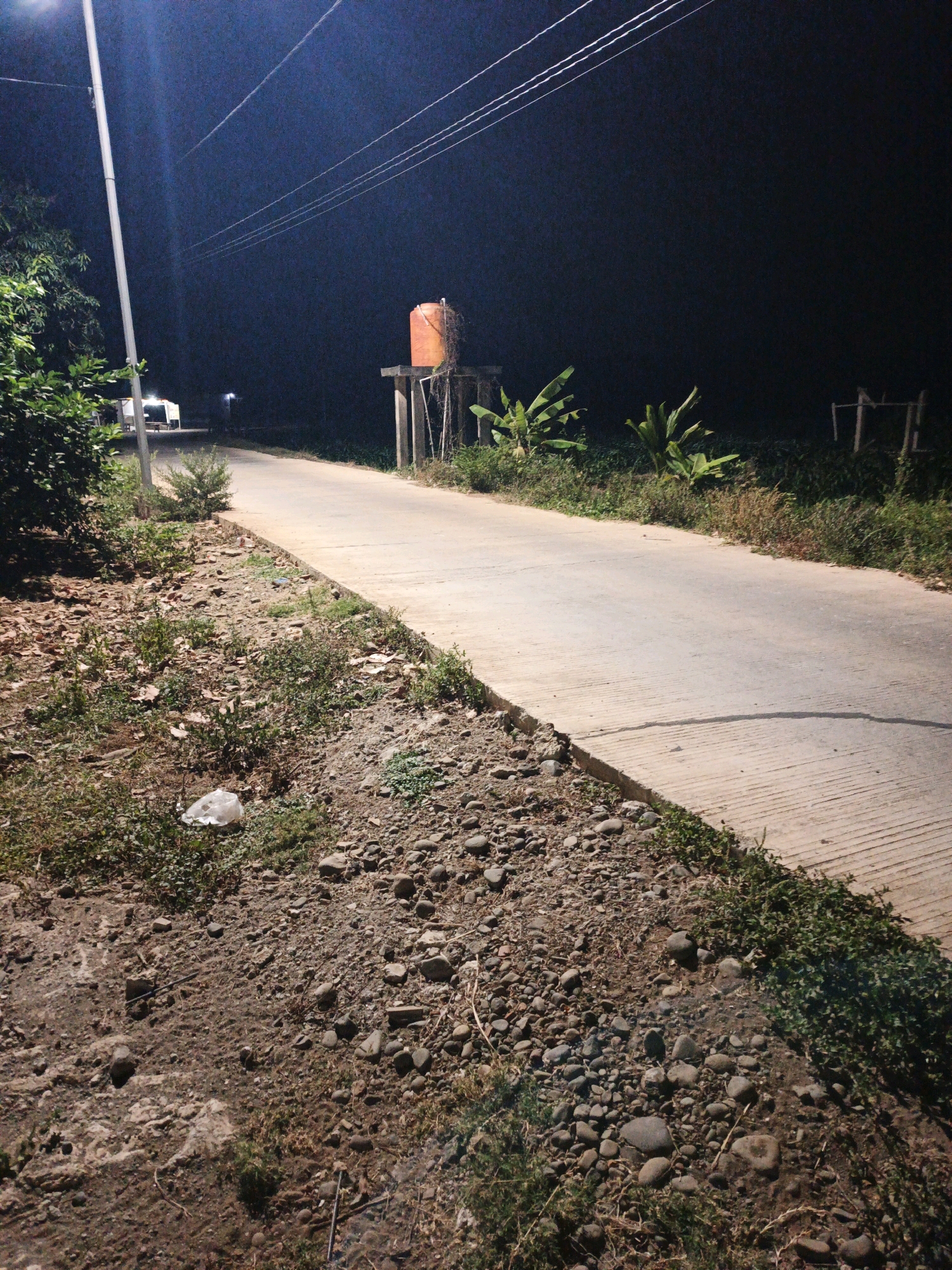 The image depicts a nighttime scene on a paved road in Bumiharjo, Indonesia. To the left of the road, there is a streetlamp and dense foliage. The road itself is bordered by a gravelly and grassy shoulder, with scattered rocks and some litter, including a plastic bag. In the distance, a white vehicle is parked near a building illuminated by a light. Further along the road, an orange water tank stands on a raised platform supported by concrete pillars. To the right of the water tank, banana plants are visible, and beyond them, the landscape fades into darkness. Power lines stretch across the dark night sky. The scene is captured from a low angle, emphasizing the road and its immediate surroundings.