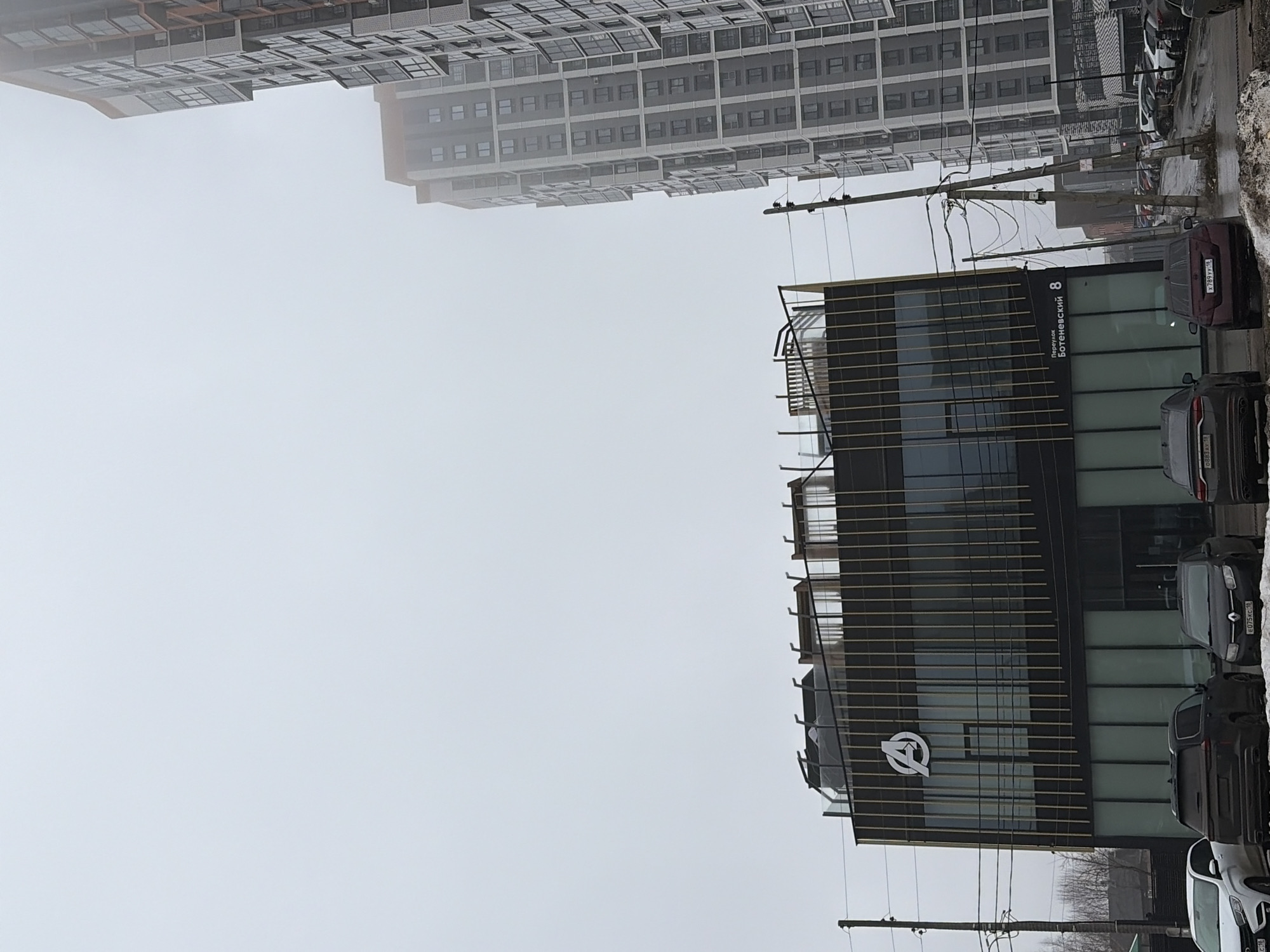 The image shows a street scene in Izhevsk, Russia, under overcast conditions. Several multi-story apartment buildings dominate the background. A modern building with a distinctive architectural style stands prominently to the right, adorned with a logo resembling the Avengers symbol. Signage on the building indicates the address as "8, Perekok Botenevskii". Several parked cars are visible along the street adjacent to the building. Utility poles and wires are strung across the sky.