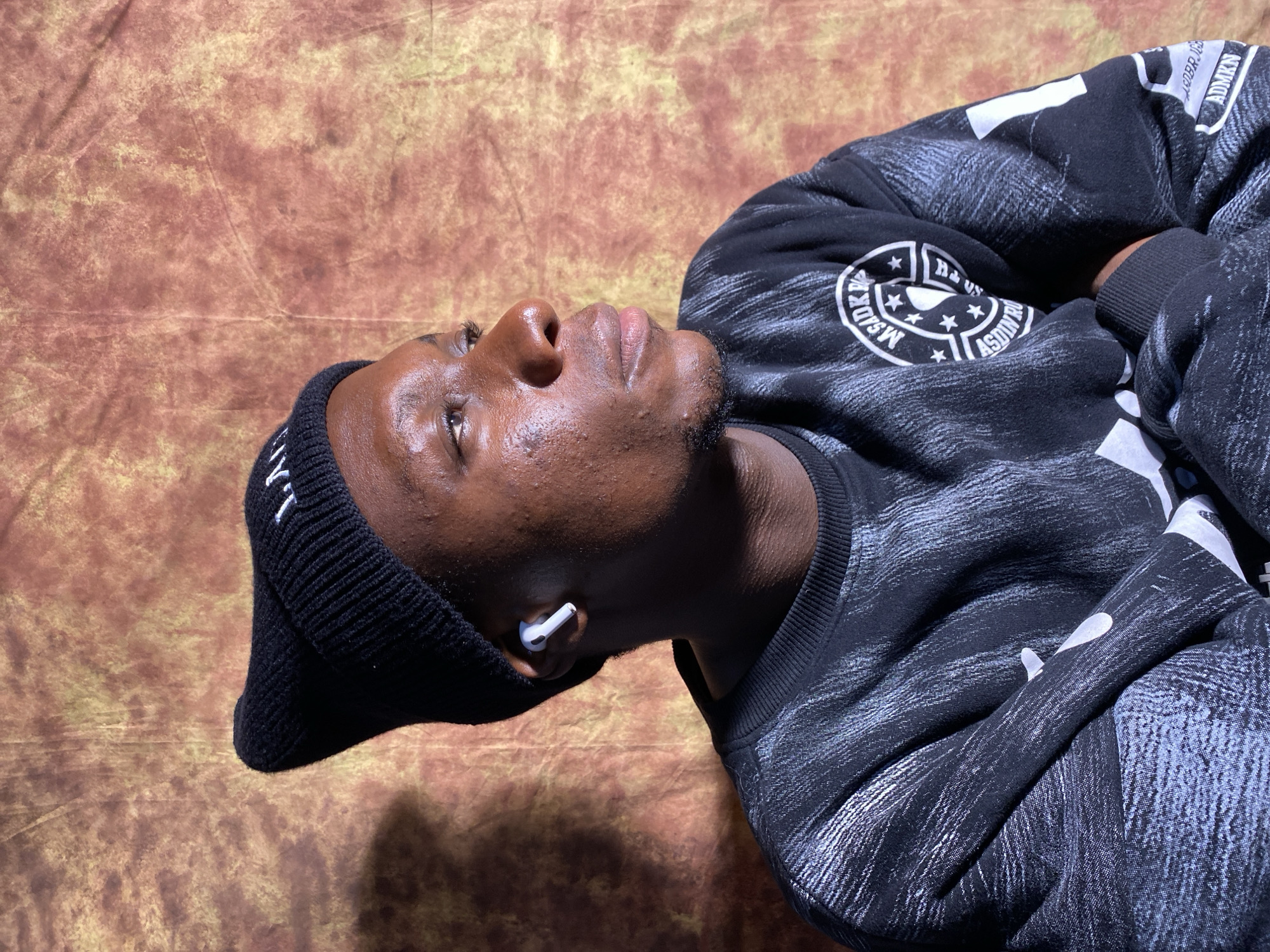 A headshot of a young Black male wearing a black beanie and a black sweatshirt with a distressed pattern. He is looking to his right, with a white earbud in his right ear. The background is a textured, mottled brown and yellow cloth. The image is lit from the left, casting a shadow on the right side of his face. There is no indication of the city of Barakin Akawo, Nigeria in the image.