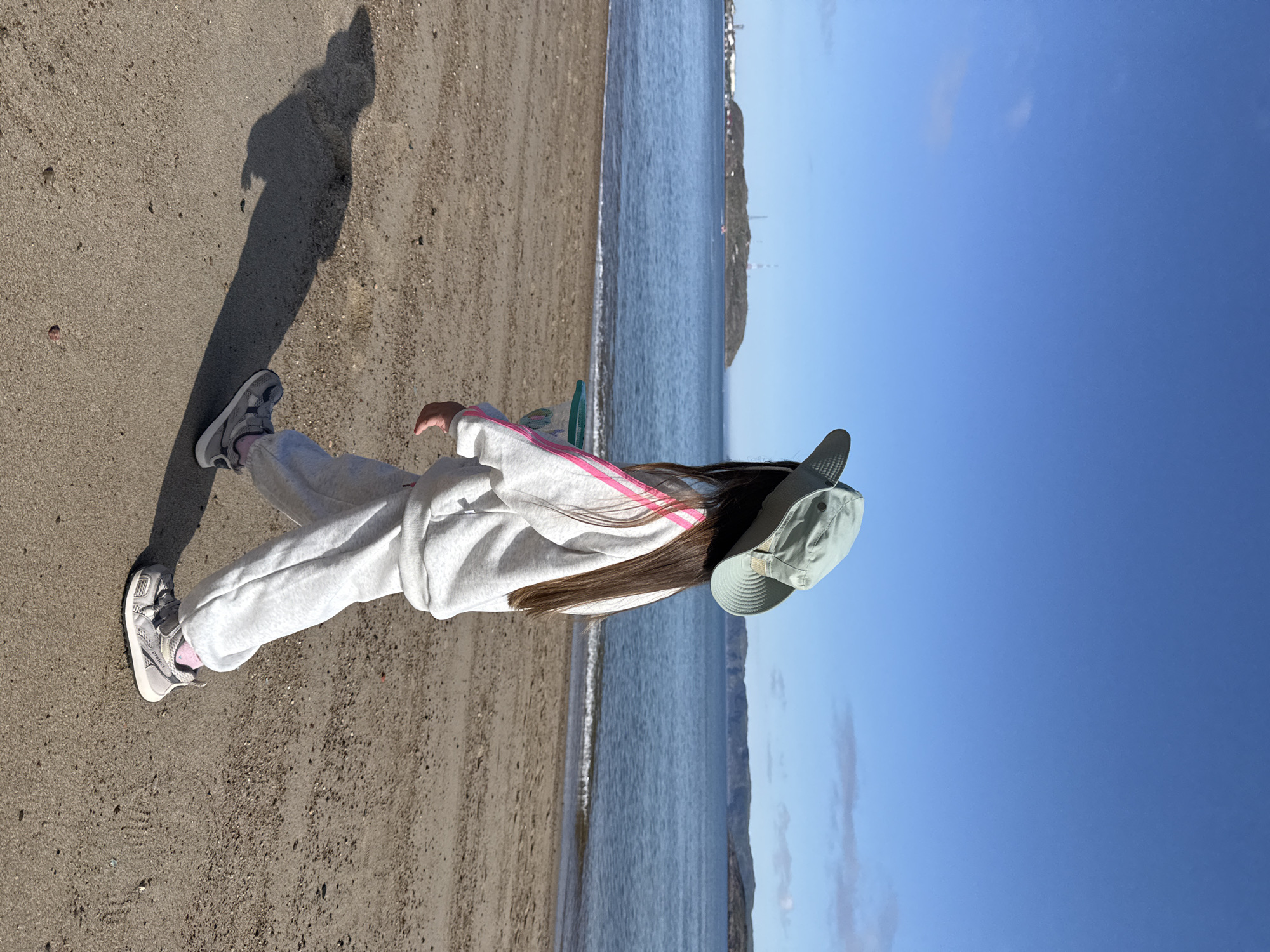 A young girl, facing away from the camera and wearing a light green wide-brimmed hat, a grey tracksuit with pink stripes on the sleeves and pants, and grey sneakers with pink accents, walks on a sandy beach. She is holding a green and white speckled net with a handle. The beach is composed of coarse sand and small pebbles, and her shadow stretches out to her left. In the background, a calm, blue body of water meets the shore, with gentle waves lapping at the sand. Beyond the water, a low, rocky island with some structures and communication towers is visible. The sky is clear and bright blue with a few wispy clouds. The scene appears to be set in Teriberka, Russia, a coastal settlement known for its dramatic Arctic landscapes.
