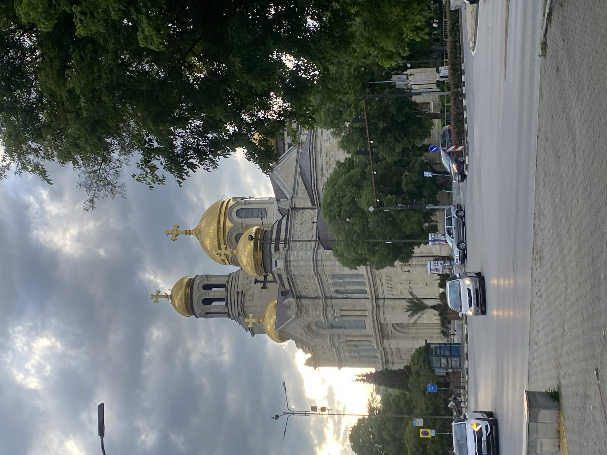 The image shows the Cathedral of the Assumption of the Virgin in Varna, Bulgaria. The cathedral, with its gold domes and intricate facade, dominates the left side of the frame. A street runs along the right side, with trees lining both sides of the road. Several cars are present on the road. The sky is cloudy, with some sunlight visible behind the cathedral. A street lamp is visible on the far left. The image is taken from a slightly elevated perspective. The photo is rotated to the side.