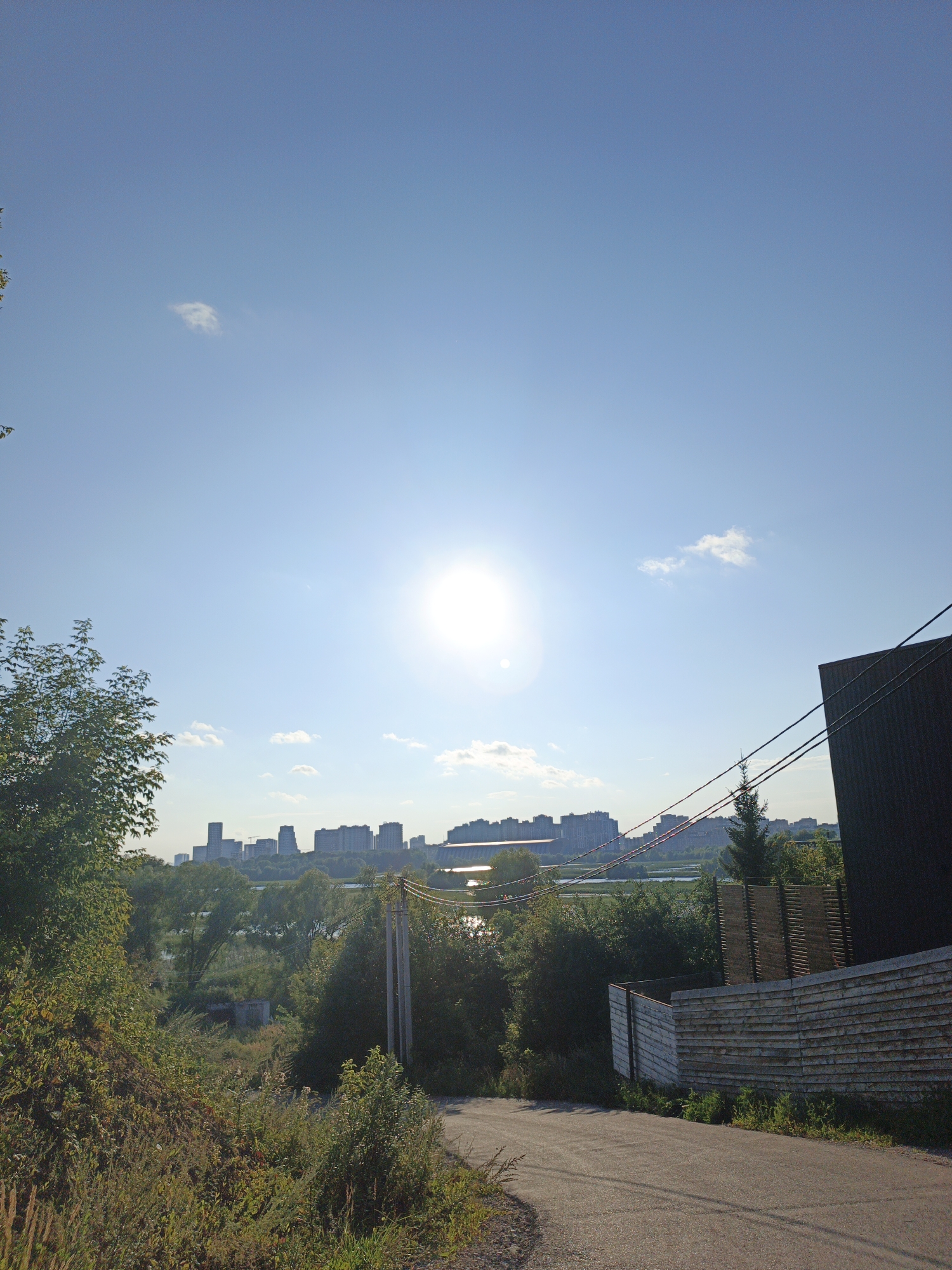 A sunny, clear sky dominates the upper portion of the image, with the sun prominently visible as a bright orb slightly left of center. The lower half of the image depicts a semi-urban landscape. In the foreground, a paved road curves from the bottom right towards the center. To the right of the road is a dark, modern building with a sloped roof, and a fence made of horizontal wooden planks. Vegetation, including trees and bushes, lines the left side of the road and extends into the mid-ground. In the distance, across a body of water, a skyline of multi-story buildings is visible. The overall scene suggests a clear day in a location that blends natural elements with urban development. Based on the architecture of the distant buildings and the general appearance of the landscape, this scene is likely in Kazan, Russia.