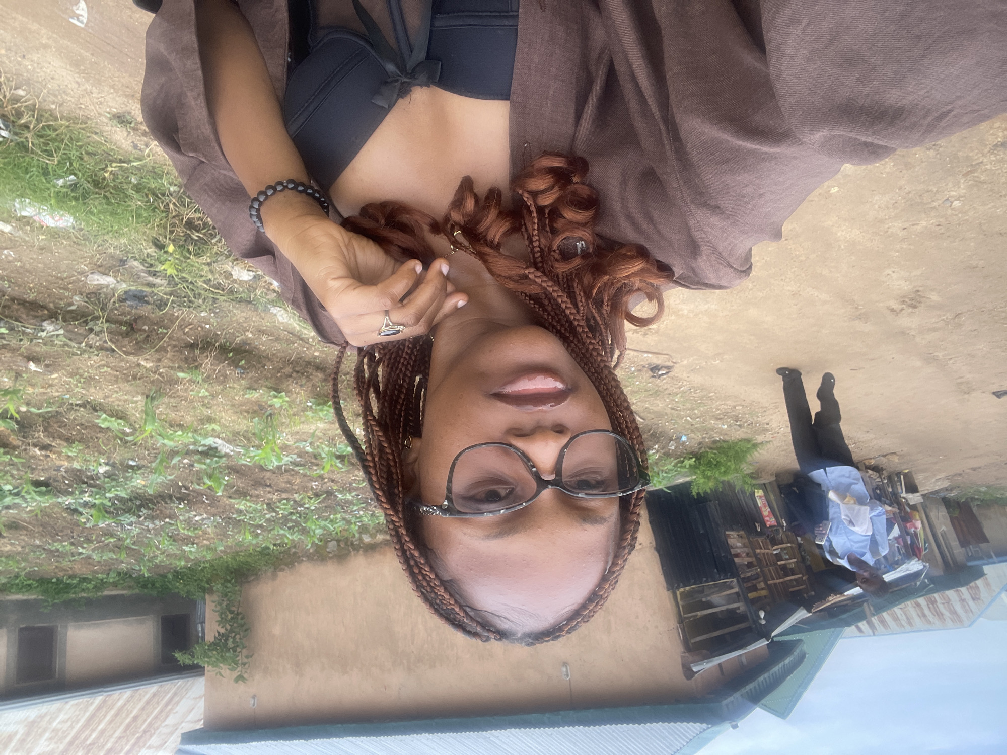 In Rafin Par, Nigeria, a young woman with braided reddish-brown hair and glasses takes a selfie outdoors. She wears a brown overshirt, a black bra, and a black bracelet, with a ring on her left hand. In the background, a man in a light blue shirt and dark pants walks away from the camera, holding a white cloth and a phone. Buildings with corrugated metal roofs and a dirt path are visible. A small field with young corn plants and scattered debris is to the right. The sky is overcast.