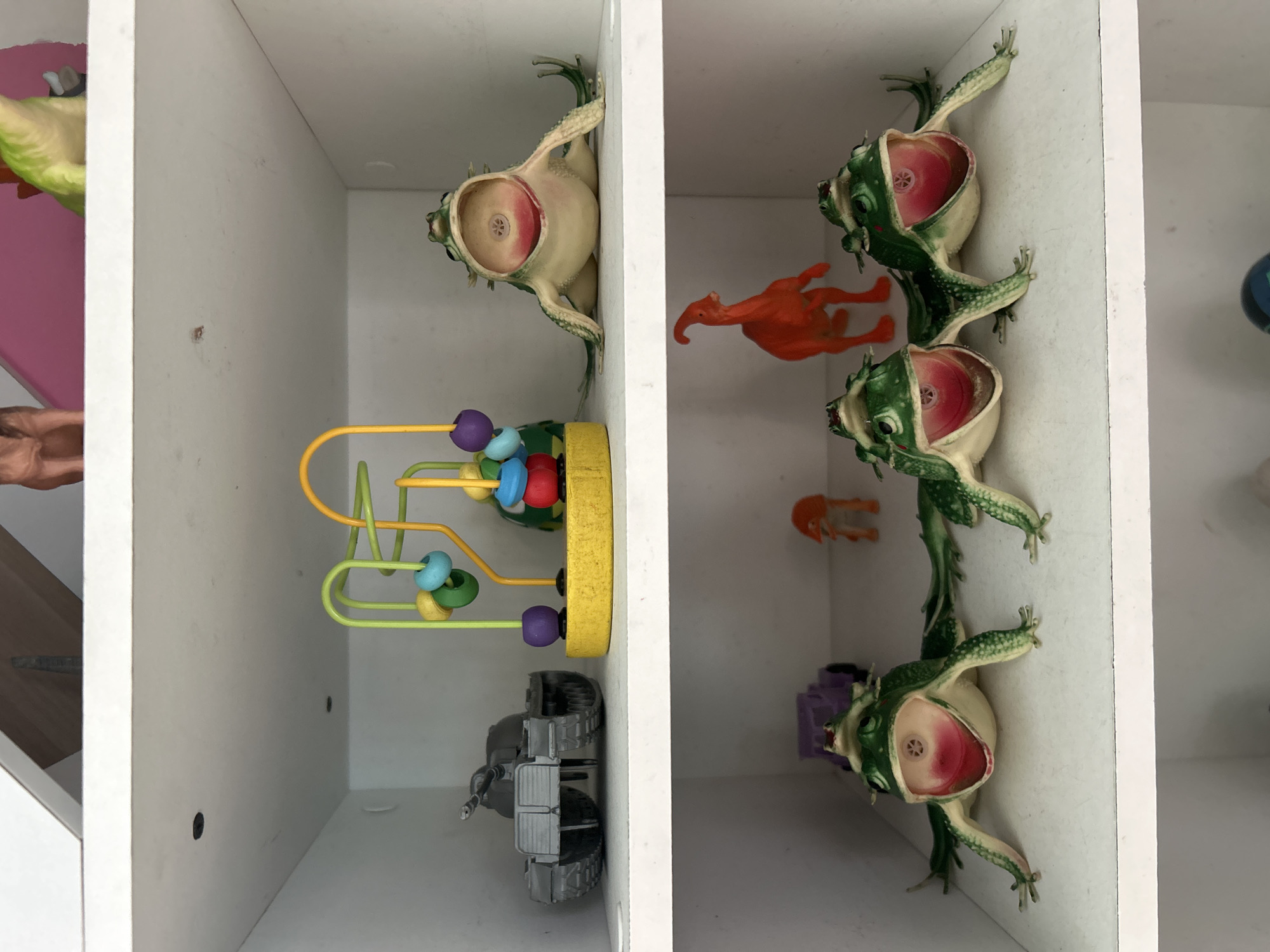 The image shows two shelves of a white cabinet filled with various toys. On the top shelf, from left to right, are a gray toy tank, a colorful bead maze toy, and a green toy frog with its mouth open. On the bottom shelf, from left to right, are a purple toy truck, three green toy frogs with their mouths open, and a small orange dinosaur toy. The scene is indoors, likely in a home or a toy store. There is no indication of the city of Kuta, Indonesia, or any specific location.