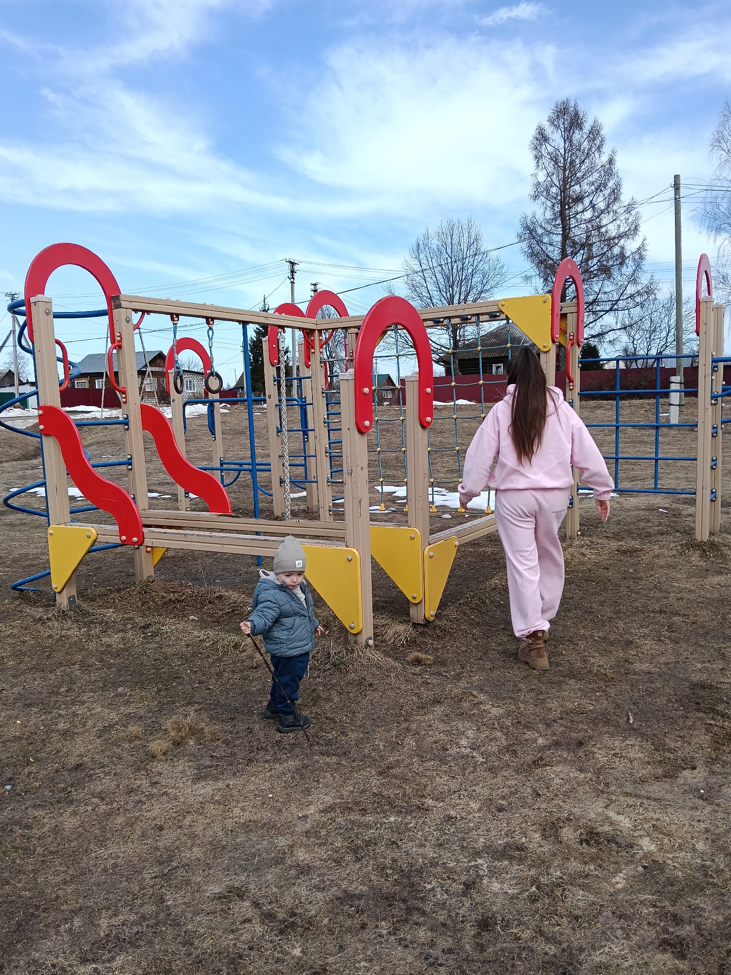 The image depicts a playground scene in Красный Маяк, Russia. A young child, wearing a gray hat and blue jacket, stands near a playground structure, holding a stick. The playground equipment features red, yellow, and blue elements, including slides and climbing structures. An adult female, clad in a pink tracksuit and brown boots, walks away from the camera. The ground cover appears to be dry grass with remnants of snow. In the background, houses and bare trees are visible under a partly cloudy sky.