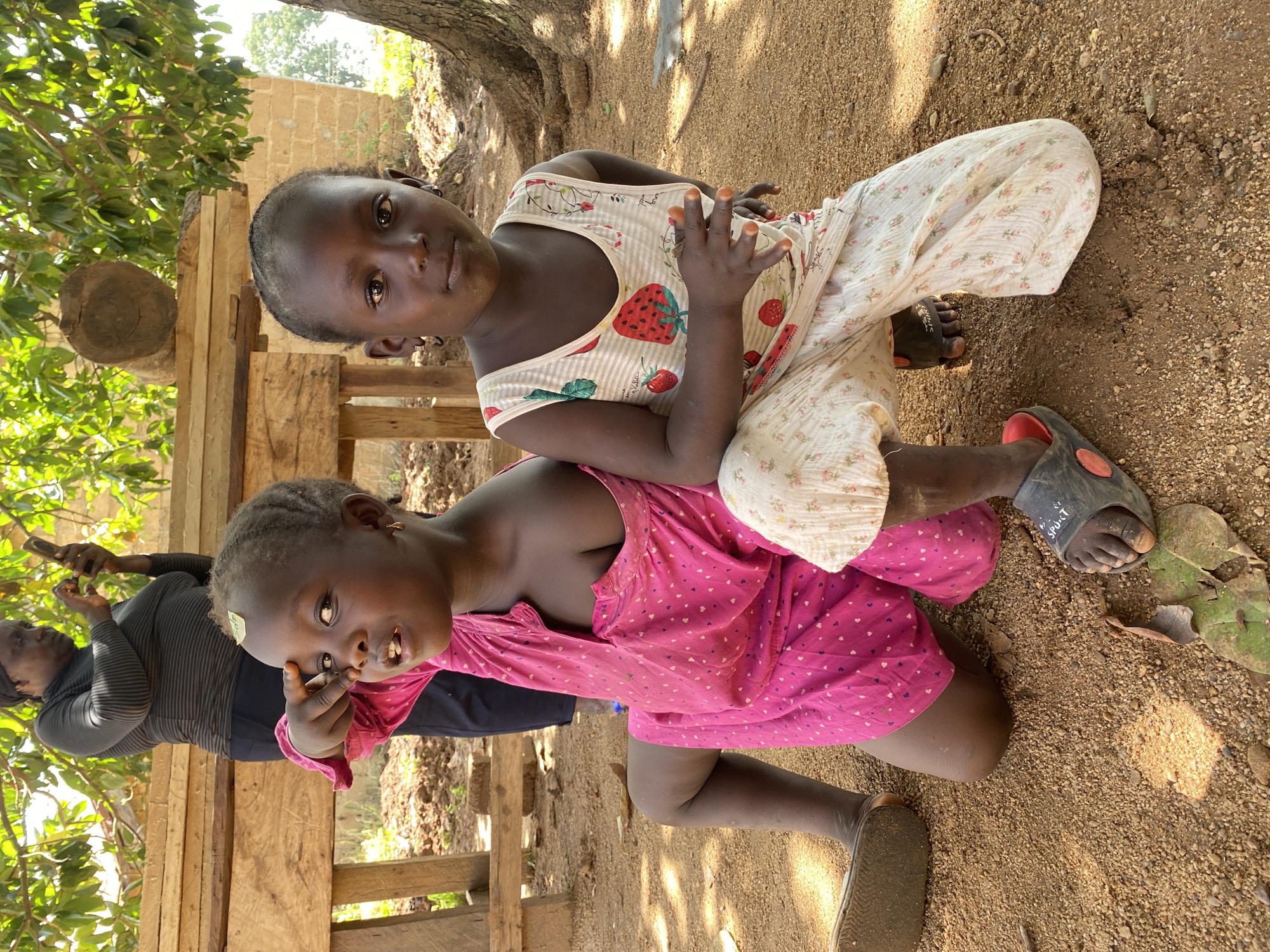 Two young girls are outdoors in Hoss, Nigeria. One girl wears a pink dress with heart patterns and sits on her knees, while the other sits on her back wearing a white strawberry-patterned top and floral pants. Both wear sandals. An adult woman stands in the background, holding a phone. The ground is dirt and gravel, with foliage and a wooden structure nearby. A tree provides shade.