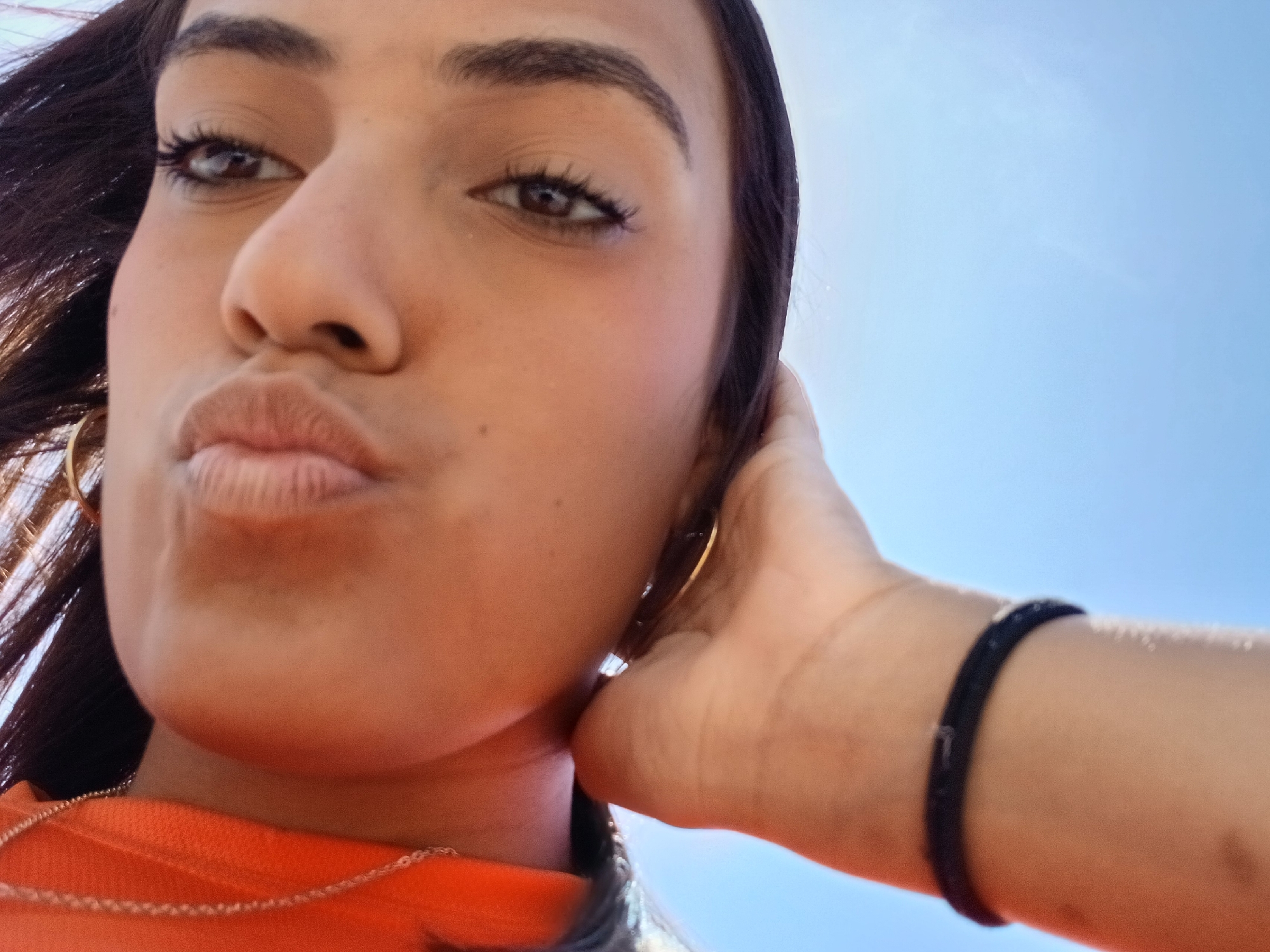 A young woman is taking a selfie. Her face fills most of the frame, angled from below. She is making a kissing face. She has dark hair, brown eyes, and is wearing gold hoop earrings and a gold necklace. She is wearing an orange shirt. Her right hand is touching the side of her face. She has a black bracelet on her right wrist. The background is a bright blue sky. There is no indication of the city of Altagracia, Venezuela.