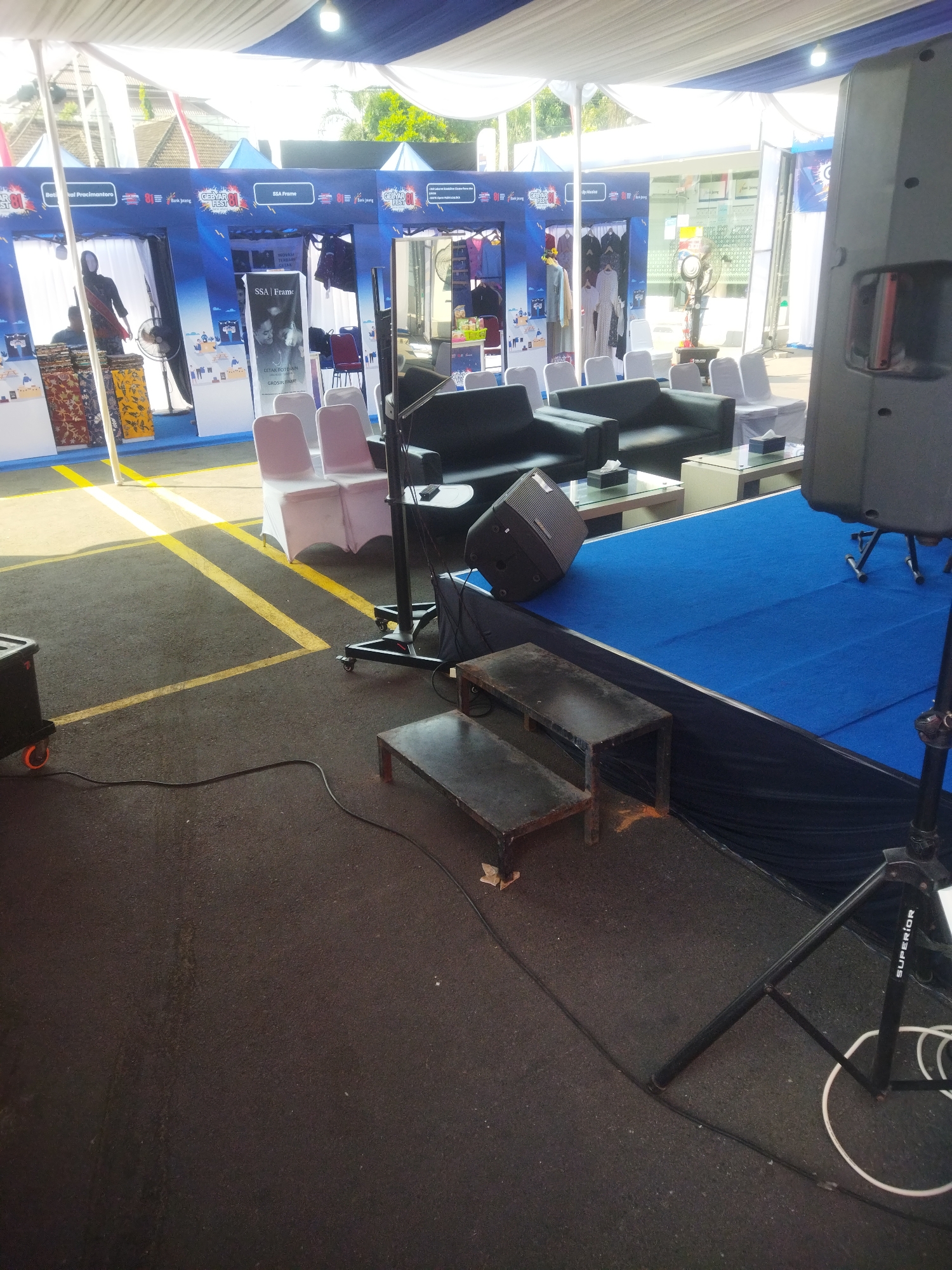 The image depicts an outdoor event setup under a white and blue tent, likely in Giripurwo, Indonesia. The scene is set up for an event with several booths visible in the background, featuring banners with Indonesian text and logos. In the foreground, there is a stage area covered with blue carpet. On the stage, there are black leather sofas and glass-top coffee tables with tissue boxes. To the right, a large speaker stands on a tripod. In front of the stage, two metal benches are placed on the ground. The ground surface is asphalt with yellow lines marking areas. The overall impression is of a temporary event space prepared for an audience.