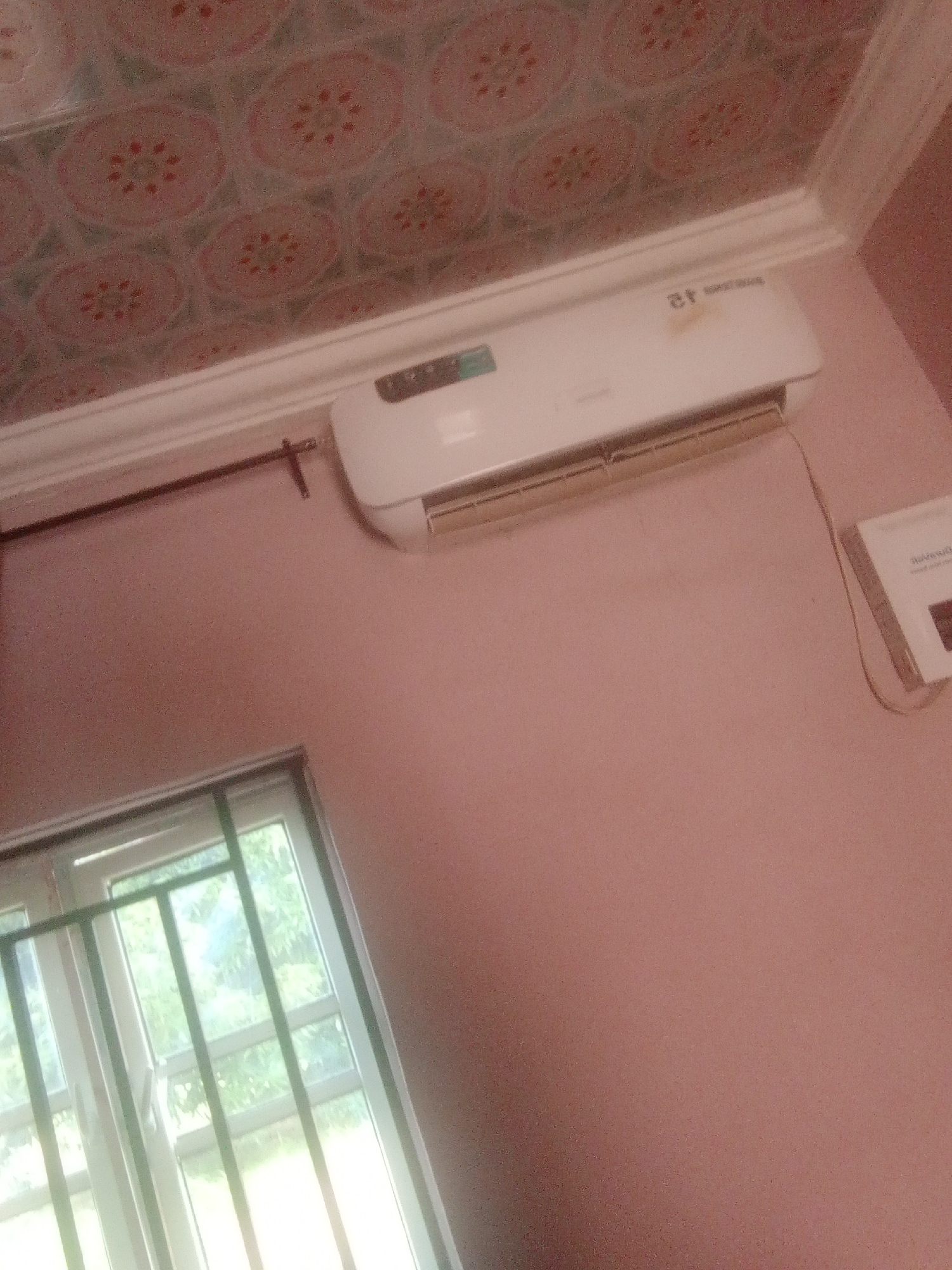 The image shows a room with light pink walls and a patterned ceiling. An air conditioner is mounted high on the wall, with a white casing and a visible vent. Below the air conditioner, a dark rod extends horizontally across the wall, attached to two supports. To the left of the air conditioner, a window with green bars is visible, with light filtering through it, suggesting it overlooks an outdoor area with foliage. A white object with some text on it is mounted on the wall to the right of the air conditioner, with a thin wire connecting it to the air conditioner. The image appears to be taken indoors in Jalingo, Nigeria.